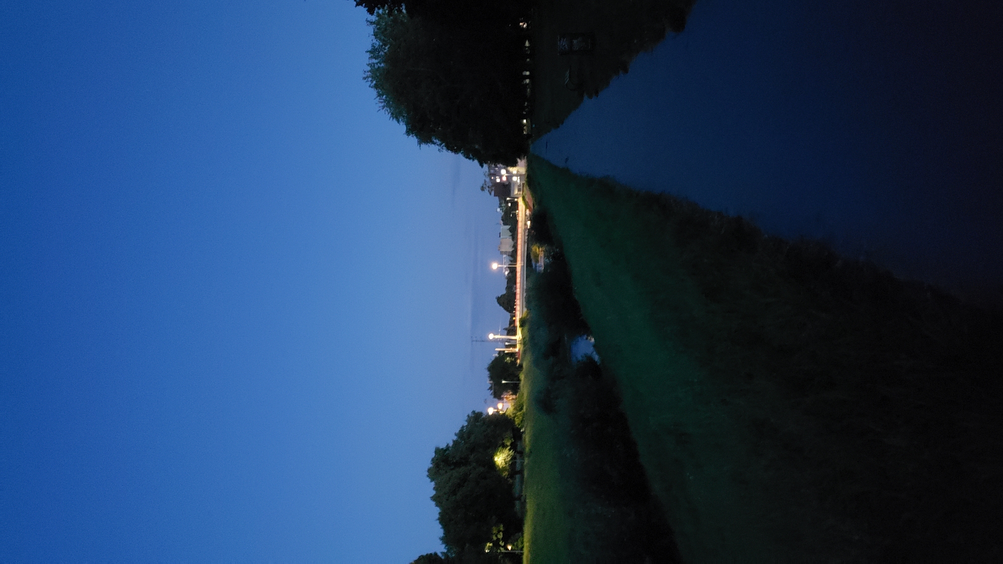 The image depicts a nighttime scene of a park or green space in Prešov, Slovakia, likely during twilight or shortly after sunset, given the deep blue sky. A paved pathway runs along the right side of the frame, receding into the distance. To the left of the pathway, a grassy embankment slopes down towards a narrow body of water, possibly a stream or small river, which reflects the dark blue of the sky.
On the far side of the water, a row of trees with visible lighting from street lamps can be seen. Further in the background, a bridge with illuminated railings spans the water, and beyond that, buildings and more streetlights are visible, suggesting the proximity of an urban area. The overall atmosphere is tranquil and slightly dusky. No people are present in the image.