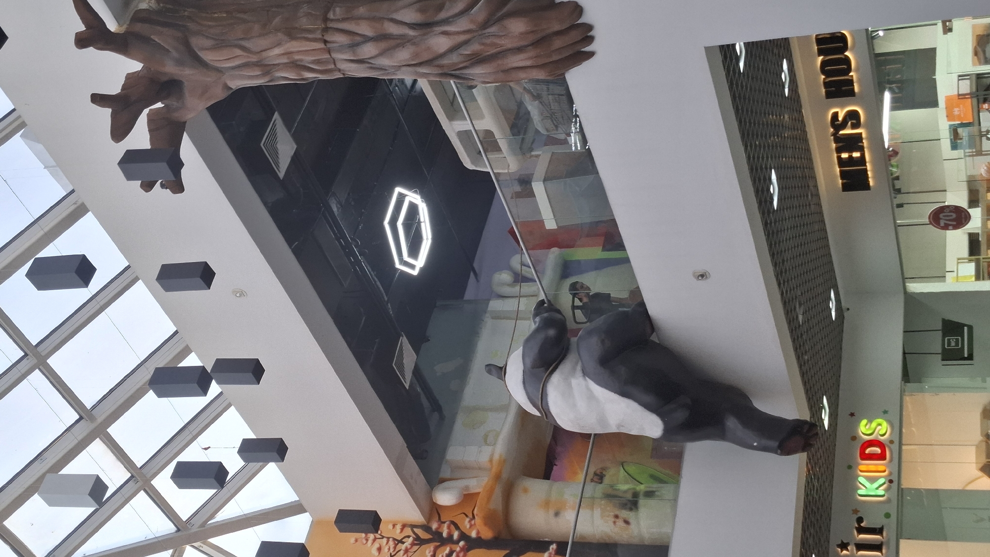The image captures an indoor scene, likely a shopping mall, with a large, stylized tree sculpture dominating the upper portion. Below the tree, a large panda statue is seen playfully hanging from a glass enclosure, its body draped over a ledge. The panda is depicted in a black and white cartoon style, with a white belly.
The setting appears to be a mall or large retail space, with various shop fronts visible in the background. One sign clearly reads "MEN'S HOUSE" in illuminated golden letters, suggesting a men's clothing store. Another sign, with colorful letters, says "KIDS," indicating a children's store. Other visible elements include geometric light fixtures on the ceiling and what appear to be decorative, rectangular objects suspended from the ceiling. A "WC" sign is also visible, pointing to restroom facilities. The overall ambiance seems bright and designed for shoppers, suggesting it's daytime or the mall is well-lit. The image is oriented sideways, with the subjects appearing upside down. There are no clear indications of weather or time of day beyond the indoor lighting. The activity taking place is static, focused on the display of the panda statue and the retail environment.