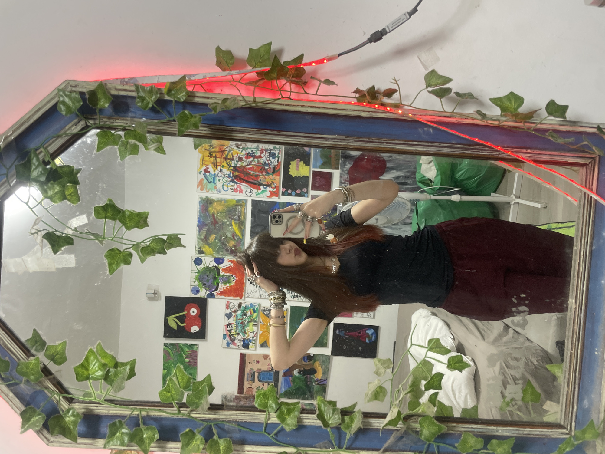 A reflected image of a woman taking a selfie is visible through a mirror. The woman has long brown hair and is wearing a black top and burgundy pants. She is holding a smartphone in her right hand, and her left hand is touching her hair. She is wearing numerous bracelets on her right arm and rings on both hands.
The mirror frame is decorated with artificial ivy and red LED lights. The wall behind the mirror is covered with small paintings and drawings. A white fan is to the right of the mirror. The room appears to be interior, possibly a bedroom or studio.
There is no information provided to confirm that this image was taken in the city of Raanana, Israel.