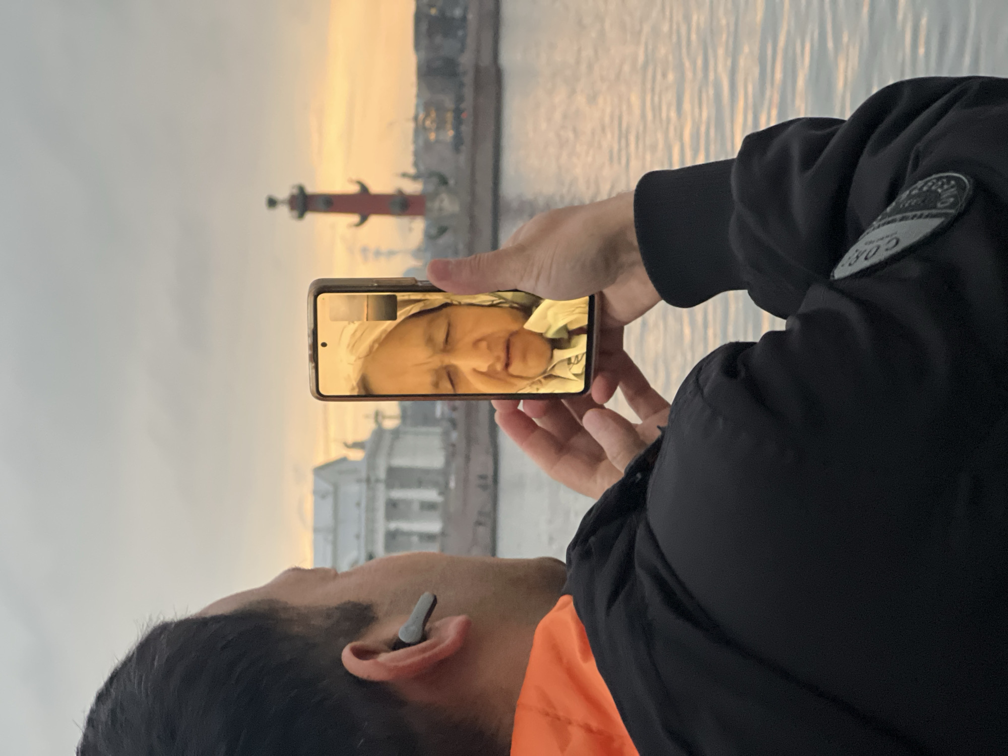 A person wearing a black jacket with an orange lining stands by the Neva River in Saint Petersburg, Russia, holding a smartphone in landscape mode. The smartphone's screen displays a video call of a woman in what seems to be a bedroom or hotel room, as evidenced by the bed frame. In the background, across the Neva River, are the Rostral Columns, visible in the sunset light, and the facade of the building. The sky has a mix of orange and gray hues, indicating it might be sunset or sunrise. The person taking the photo has a gray earbud in their ear.