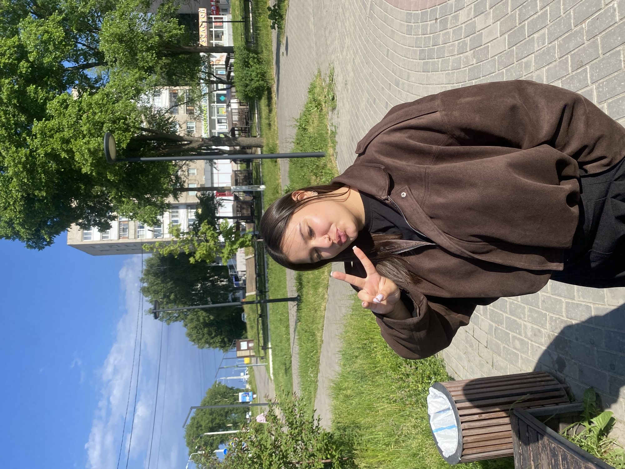 A young woman stands outdoors in Chaikovsky, Russia, on a sunny day. She wears a brown jacket and is making a peace sign with her right hand while puckering her lips for a kiss-like expression. The background features a brick-paved area transitioning into grass, with trees, lamp posts, and a building visible in the distance. A trash can sits nearby.