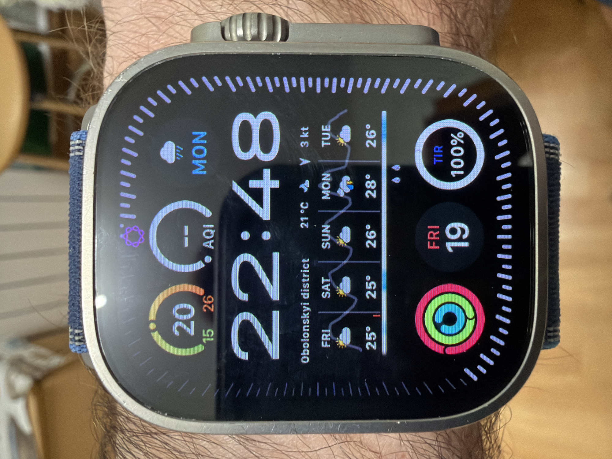 A close-up, eye-level shot shows a person's wrist wearing a smartwatch displaying the time as 22:48. The watch face features multiple complications, including the current temperature, weather icons, and daily activity rings. The background consists of blurred indoor elements, suggesting the photo was taken in a domestic setting. The watch indicates a location of "Obolonskyi district" in Kyiv, Ukraine, as it displays weather forecasts for Friday, Saturday, Sunday, Monday, and Tuesday with temperatures ranging from 25 to 28 degrees Celsius. A circular graphic with three colored rings, likely representing activity goals, is visible on the left side of the watch face. To the right, another circular graphic displays "TIR 100%", possibly indicating a health or fitness metric. The overall impression is a functional and data-rich smartwatch interface.