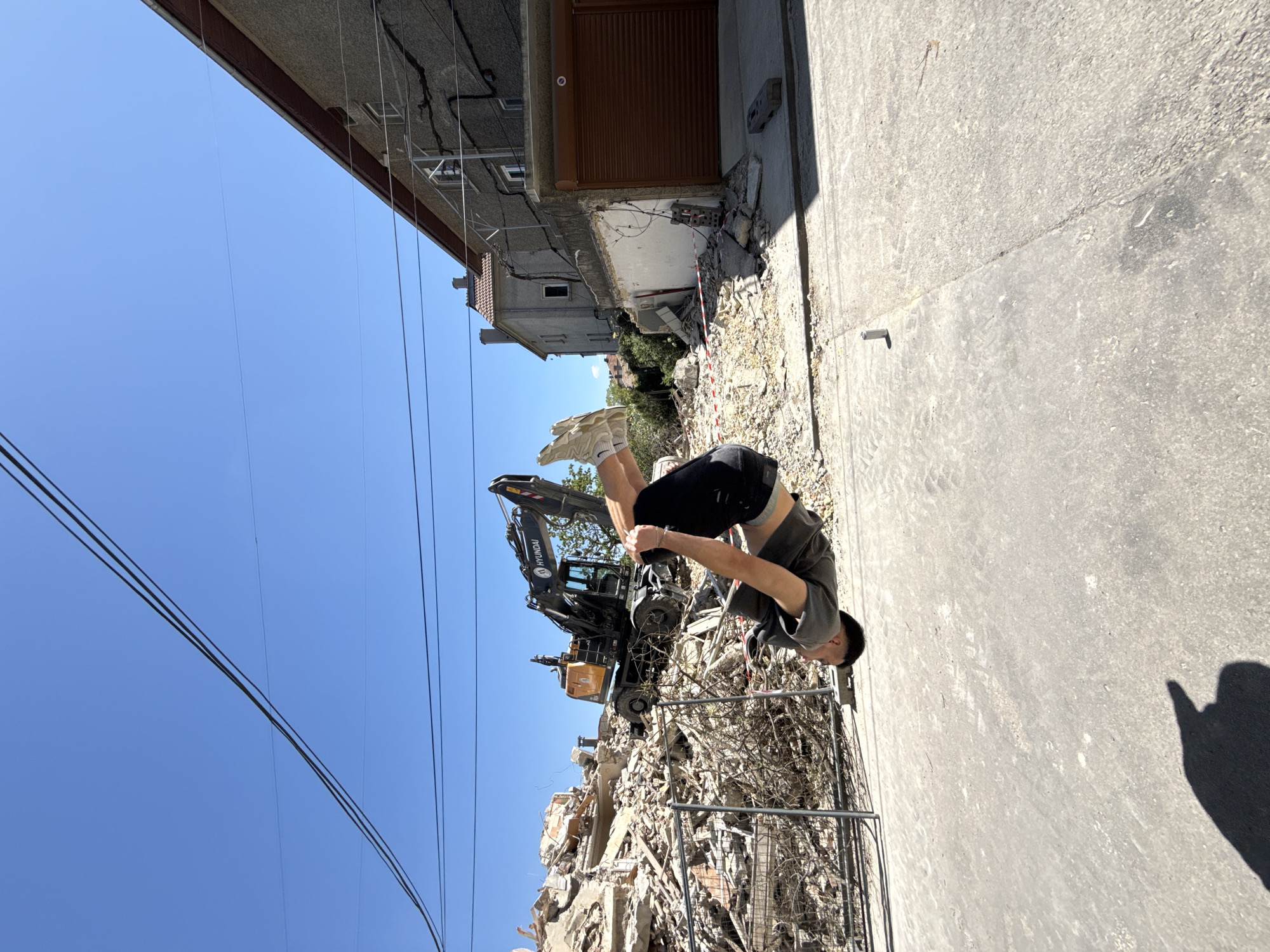 The image shows a man performing a backflip on a street in Shumen, Bulgaria. Behind him is a pile of rubble and debris, likely from a demolished building. An excavator is parked atop the rubble. Overhead are power lines. The buildings in the background are of older construction, and some appear damaged. The sky is clear and blue. The street is concrete and in relatively good condition. A metal fence separates the street from the rubble pile.