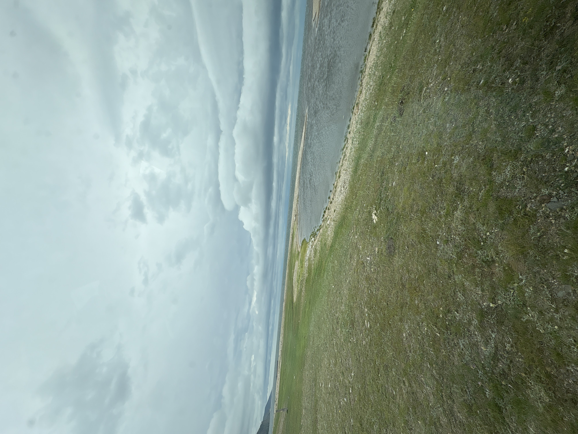 The image shows a wide, slightly choppy body of water, likely a lake, under a heavily overcast sky. The water stretches towards a distant, low-lying shoreline that appears to be lined with small stones or pebbles. To the left of the water is a gently sloping grassy embankment. The grass is a mix of green and brown, indicating a natural, unmanicured area. In the distance, a faint, darker shape suggests a mountain or hill on the far side of the lake. The sky is filled with large, grey clouds that obscure any direct sunlight. The overall impression is one of a cool, possibly windy, day in a natural, remote setting. The image does not contain any people or specific man-made structures that would allow for precise identification of the location beyond the general environmental description.