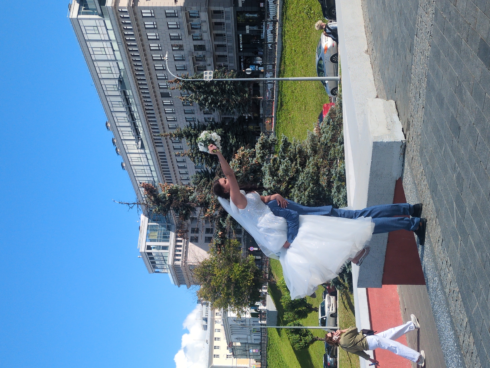 This media file captures a candid moment during what appears to be a wedding celebration. The primary subjects are a bride, dressed in a sparkling white wedding gown and veil, and her partner, wearing blue jeans and a jacket. The bride is holding a bouquet of white flowers aloft and appears to be posing playfully, with her partner supporting her. They are standing on a paved area, possibly a plaza or sidewalk, with a low white barrier beside them.
In the background, a modern, multi-story building with numerous windows dominates the left side of the frame. Across from the building, there is a lush green lawn and a line of trees. Beyond the greenery, more buildings are visible, some with classical architectural features. Several cars are parked along the street. The sky is a clear, bright blue, suggesting it is daytime, likely with good weather. The sunlight casts shadows, indicating a sunny day.
A second woman, dressed in casual clothing (olive green shirt and white pants), is also visible on the paved area in the foreground, looking at her phone and holding a selfie stick, suggesting she might be taking photos or videos of the event or the couple. The overall scene conveys a sense of joy and celebration. There is no visible text within the image. The context provided indicates the location as Nizhny Novgorod, Russia.