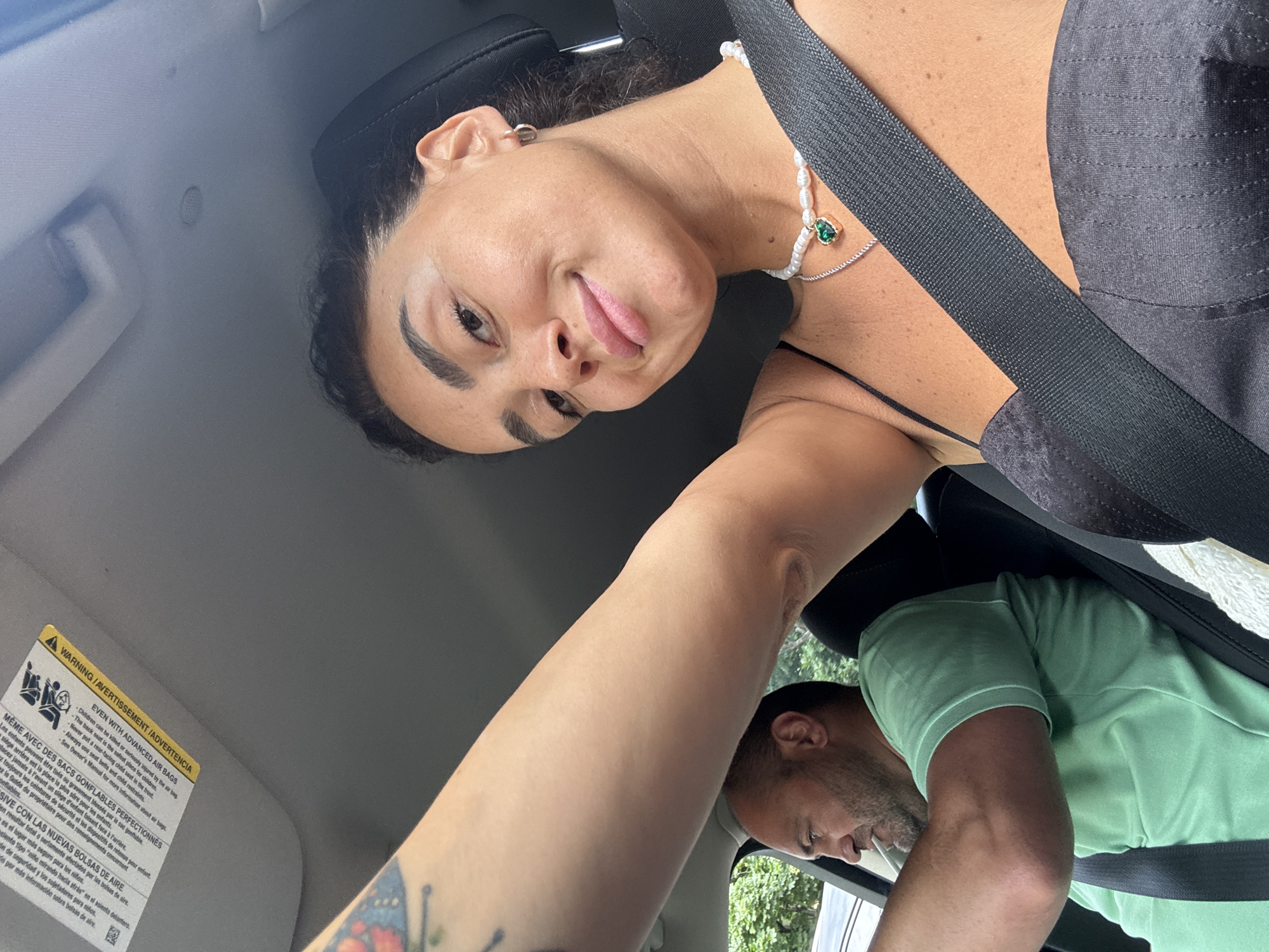 A selfie taken from the passenger seat of a car shows a woman with dark hair, wearing a black tank top and a pearl necklace with a green heart pendant, looking at the camera and smiling. Her right arm is extended, holding the camera. In the driver's seat, a man with short dark hair and a goatee, wearing a light green t-shirt, is looking to his left. The interior of the car, including a seatbelt, the dashboard, and the sun visor, is visible. The exterior of the car shows trees and foliage, suggesting an outdoor setting. The sun visor has warning labels in multiple languages, including English and Spanish, indicating a vehicle manufactured for sale in international markets. This scene does not provide any specific details to identify the location as Tbilisi, Georgia.