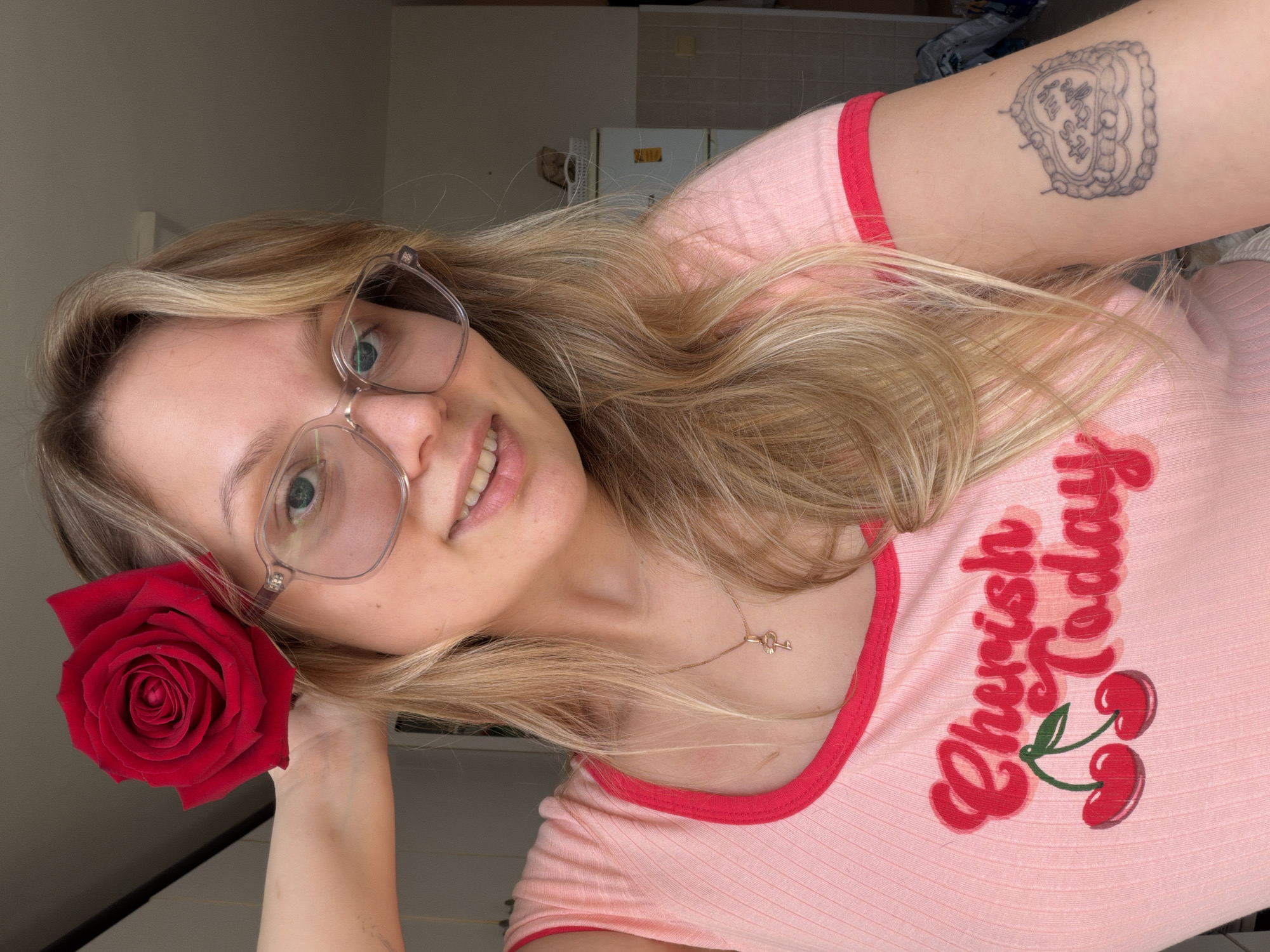 A close-up, tilted portrait features a young woman with long, flowing blonde hair and clear-framed glasses. Her light-colored eyes, possibly blue or green, gaze towards the viewer through the lenses, and a gentle smile plays on her lips. She holds a vibrant red rose in her left hand, placed near her cheek and ear.
She is dressed in a light pink t-shirt with red trim on the collar and sleeves. Emblazoned on the shirt is the text "Cherish Today" in a red, stylized script font, alongside an illustration of two red cherries with a green stem. A delicate gold-colored necklace with a small key pendant rests on her collarbone. On her right upper arm, a heart-shaped tattoo is visible, with the words "It's My Life" inscribed within it.
The background is softly blurred, indicating an indoor setting with indistinct light-colored walls. The lighting suggests it is daytime. The woman's expression conveys a pleasant and warm demeanor. The image is situated in Tel-Aviv, Israel.