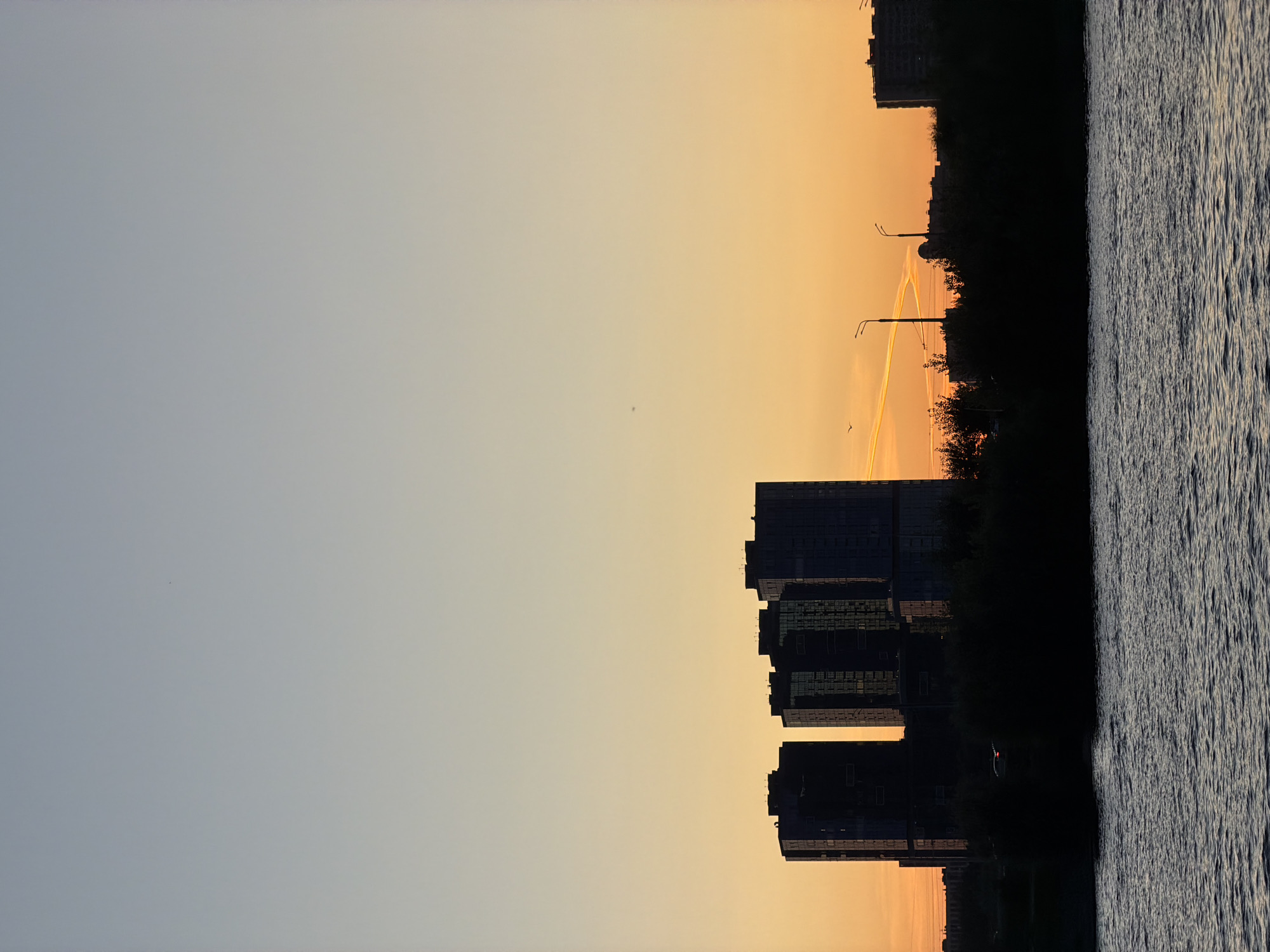 The image depicts a body of water in the foreground, likely the Kazanka River, with a silhouetted urban skyline against a vibrant sunset in the background. The sky transitions from a soft blue at the top to shades of orange and yellow near the horizon, suggesting either sunrise or sunset.
Dominating the middle ground is a cluster of modern high-rise buildings, appearing as dark silhouettes. Among these buildings, there are indications of lights within some of them. In front of the high-rises, a dark band of trees and lower structures creates a dense silhouette. Streetlights are visible above the tree line, with two distinct poles with lamps extending upwards.
Further to the right, more buildings are visible, also in silhouette, with one appearing taller than the others. There are also faint contrails in the sky, one of which glows orange from the setting sun. A single bird is visible in the sky to the left, and another is seen lower down near the contrails. A car with its taillights illuminated can be seen on a road behind the trees. The water in the foreground has ripples, indicating some movement. The overall impression is a peaceful urban scene at dusk or dawn.