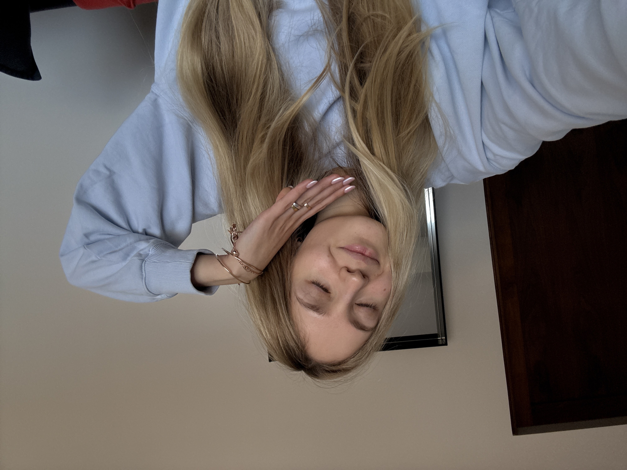 A woman with long, blonde hair, wearing a light blue sweatshirt, is seen with her eyes closed. She is wearing rings and bracelets. The background consists of a light-colored wall, and a dark wooden cabinet. The location is unknown.