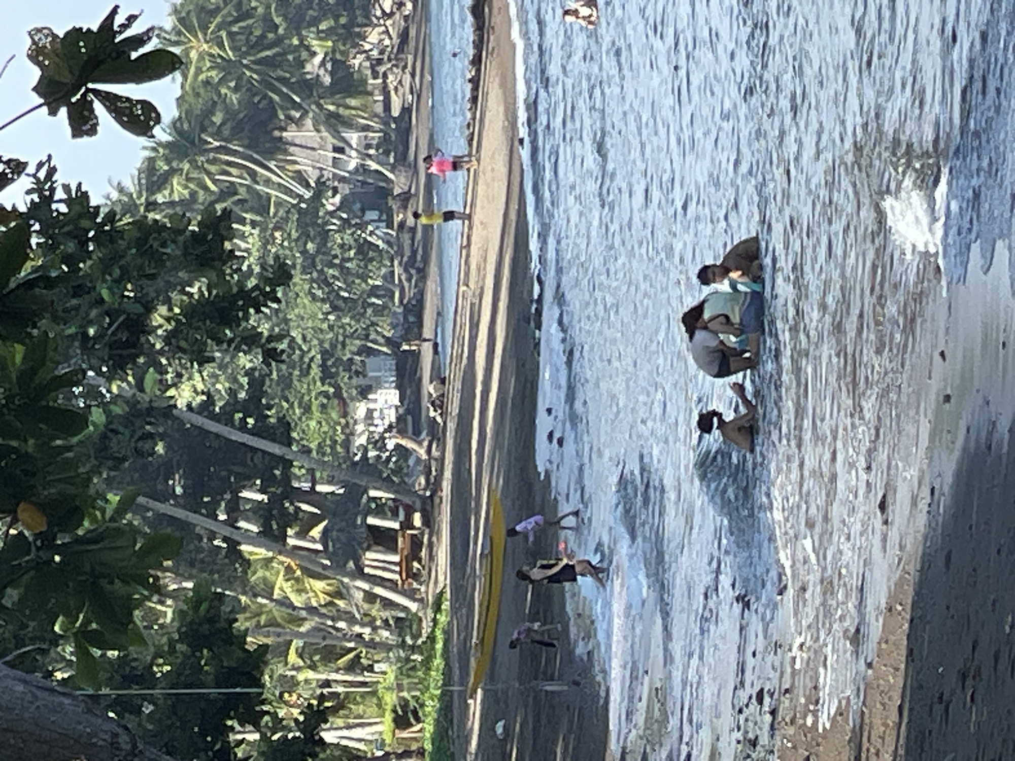 This image depicts a tropical beach scene in Dauin, Philippines. The foreground shows a shoreline with dark sand and shallow, wavy water. Several people are in the water. One group of three, two appearing to be adults and one child, are crouched together near the shore. Another person, possibly a child, is visible further out in the water to the right. In the middle ground, on the sandy beach, a yellow boat rests near the waterline. Several people are walking on the beach, including a woman in a black dress and a child being held, and two individuals wearing pink and yellow shirts. The background features a line of palm trees and some structures, suggesting a resort or residential area along the coast. Lush green foliage is prominent throughout the scene, with a clear, bright sky overhead, indicating daytime.
