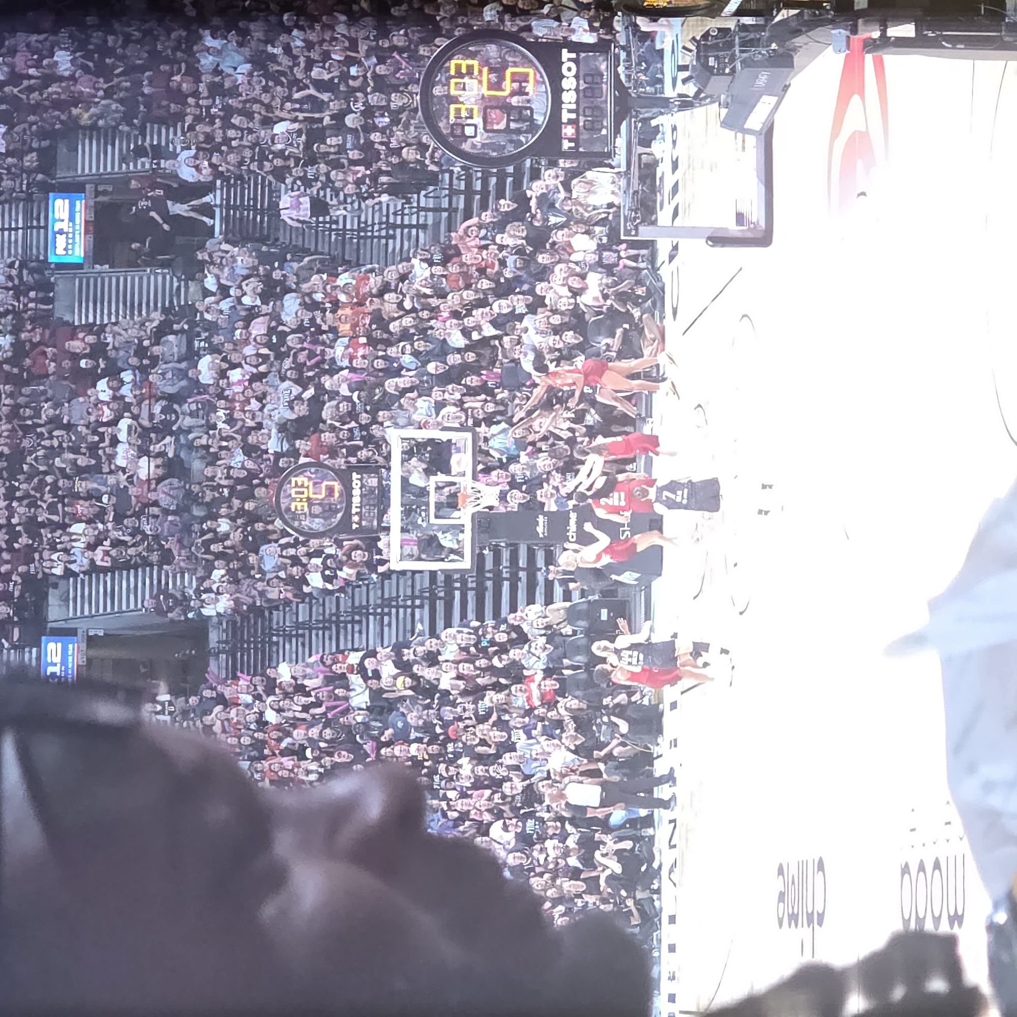 The image depicts a basketball game in progress, likely within a stadium, given the presence of a large crowd in tiered seating. The court is visible in the foreground, with players in red and black uniforms engaged in gameplay. A large circular scoreboard, featuring a Tissot advertisement, displays the time as 3:03 and the number 5. The scoreboard is positioned above a section of the crowd. In the background, a sign reads "PK12," possibly indicating a league or event. The overall lighting suggests an indoor arena setting. The presence of "Portland" signage on the court floor narrows the location to Portland, Oregon, United States of America. The perspective suggests the photo was taken from a spectator's viewpoint, with a portion of another spectator's shoulder and arm blurred in the immediate foreground, indicating it was taken from within the crowd.