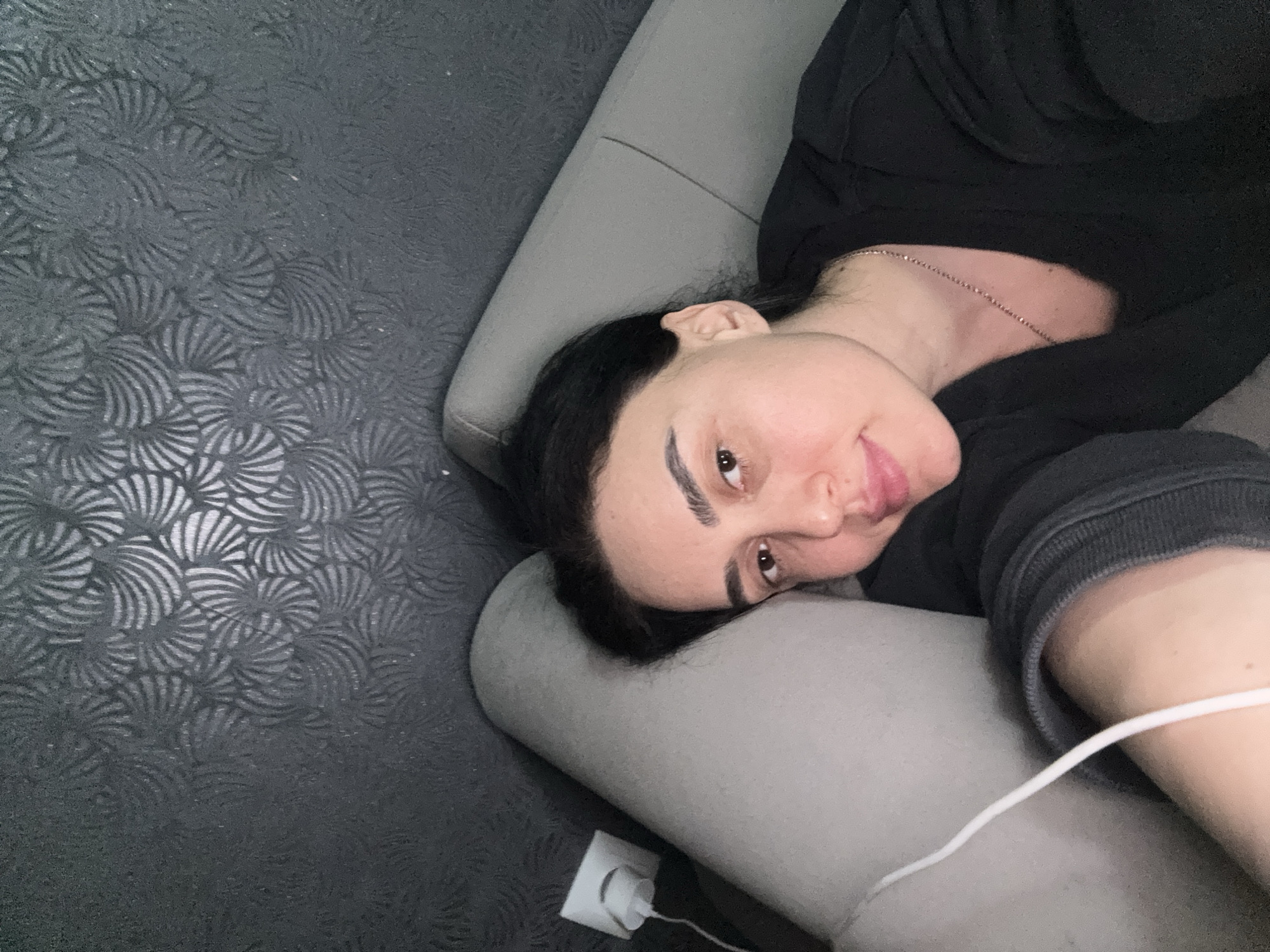A person is lying on a gray sofa against a dark, textured wall. The person has dark hair, fair skin, and is wearing a black top. A white charging cable is visible, plugged into a white wall adapter. The location is not discernible from the image.