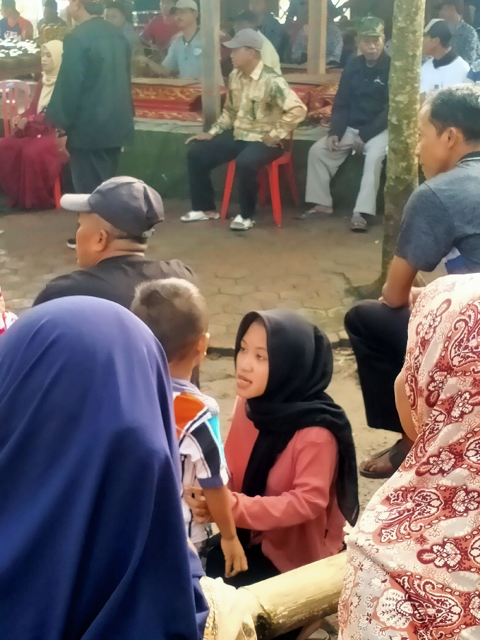 The image shows a group of people gathered in an outdoor setting. The location is in Masaran, Indonesia (latitude: -7.471, longitude: 110.957). There are at least nine people in the scene, including a young boy, a young woman, and a man with a blue hat. The people are seated in a circle, some on red plastic chairs, others on the ground. There are several trees and a wooden structure behind the people. The young woman is seated on the ground with the young boy on her left. She is wearing a pink shirt, a black headscarf, and a light pink headscarf. The young boy is wearing a colorful striped shirt. The man with the blue hat is seated on the ground, looking toward the boy and woman. The wooden structure in the background has a red roof. The floor is covered with brick and is mostly covered with sand.