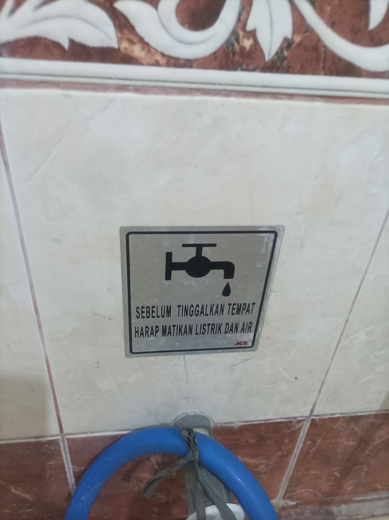 The image shows a sign with a pictogram of a faucet with a dripping droplet, mounted on a tiled wall. Below the pictogram, text in Indonesian reads "SEBELUM TINGGALKAN TEMPAT HARAP MATIKAN LISTRIK DAN AIR", which translates to "Before leaving the place, please turn off the electricity and water". A small logo, possibly indicating the manufacturer or retailer, appears at the bottom right of the sign. A blue hose is visible at the bottom of the image, partially obscuring the wall and extending out of frame. The wall features a decorative border with floral motifs above the tiled section. The overall setting appears to be a utility area or a public restroom. The image was taken in Bekasi, Indonesia.
