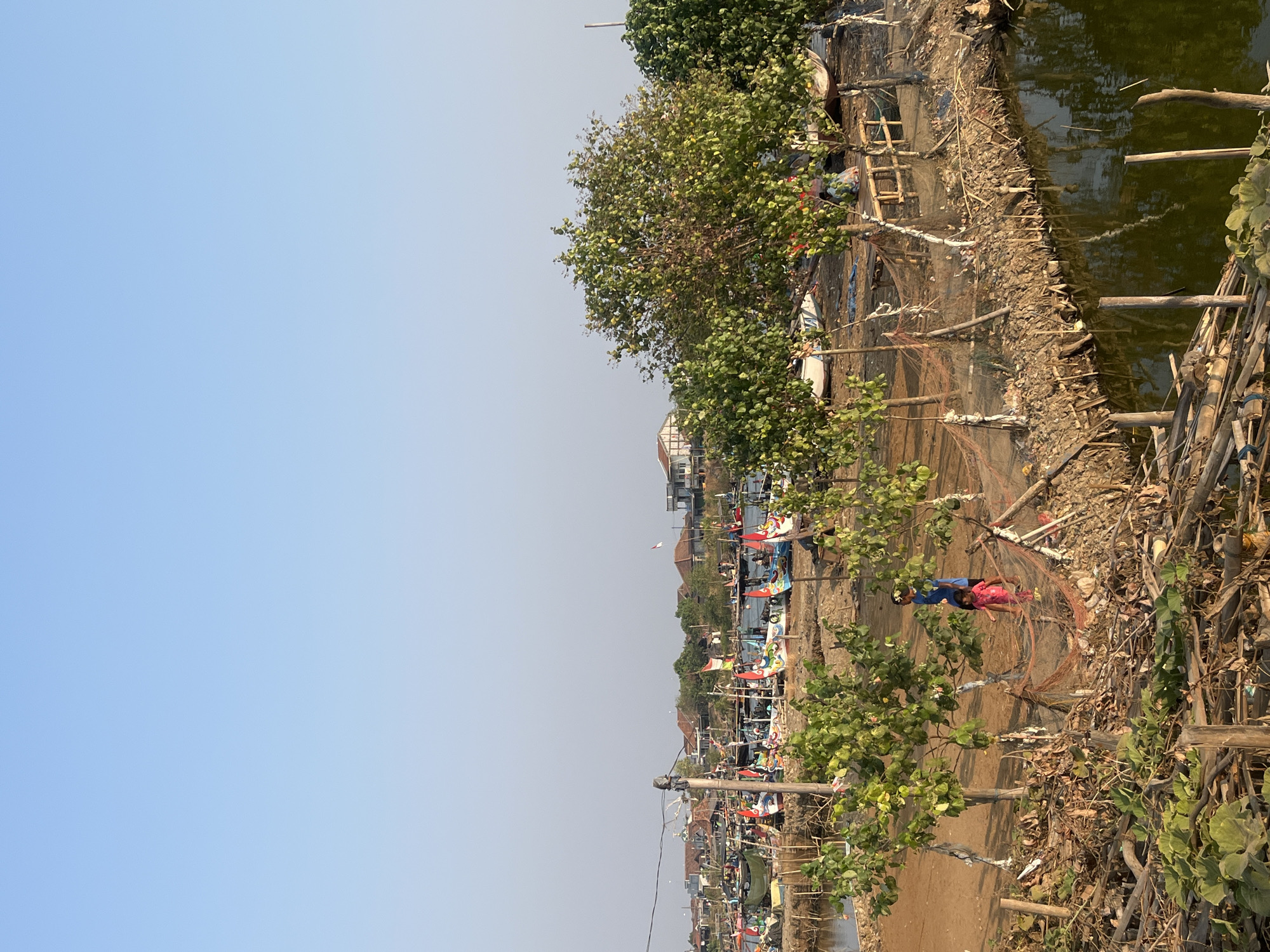 This image captures a tranquil scene in Kedungmalang, Indonesia, likely a coastal or riverside area. In the foreground, two children, one wearing a blue shirt and the other a pink dress, stand near a small tree. The ground is a mix of dirt and debris, with fishing nets draped over wooden posts. To the left and right, more trees with lush green leaves dot the landscape.
Beyond the children and trees, a body of water stretches out, reflecting the clear blue sky. Along the water's edge, numerous traditional fishing boats are moored. These boats are adorned with colorful paintings and small Indonesian flags, suggesting a festive or preparatory mood. In the background, a cluster of houses with tiled roofs and scattered trees forms a village. The overall atmosphere is peaceful and showcases a typical day in this Indonesian community, likely a fishing village.