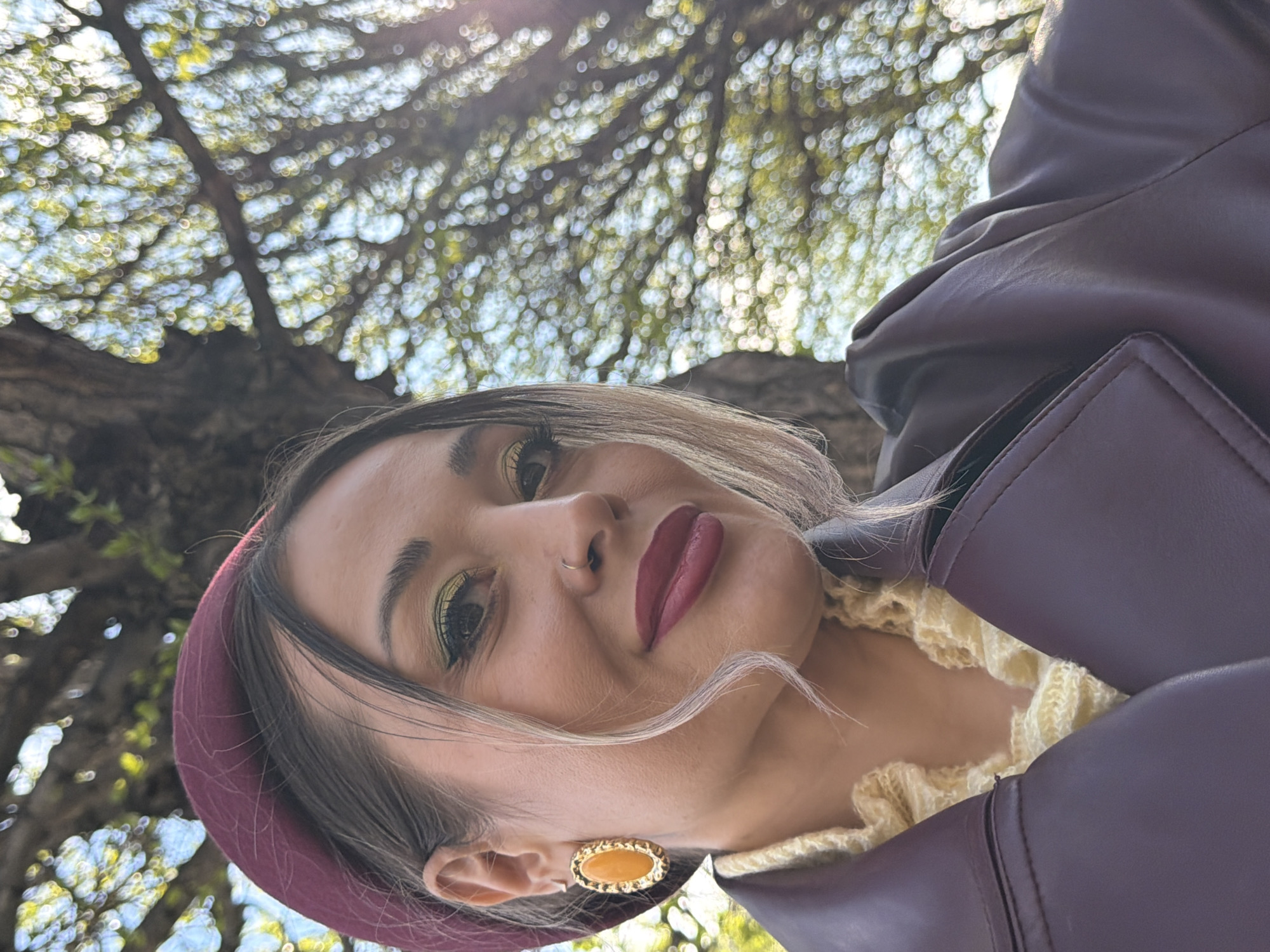 A medium, upward-angled portrait depicts a woman outdoors, her head tilted slightly to the left. She wears a maroon-colored beret, a yellow patterned sweater, and a dark burgundy leather jacket. Her makeup includes yellow eyeshadow and dark red lipstick. She has a nose ring on her left nostril and a gold-colored oval earring on her left ear. The background consists of tree branches with blurred green leaves, suggesting the location is outdoors near a tree. The exact location in the city of Krasnoyarsk, Russia, cannot be determined from the image.