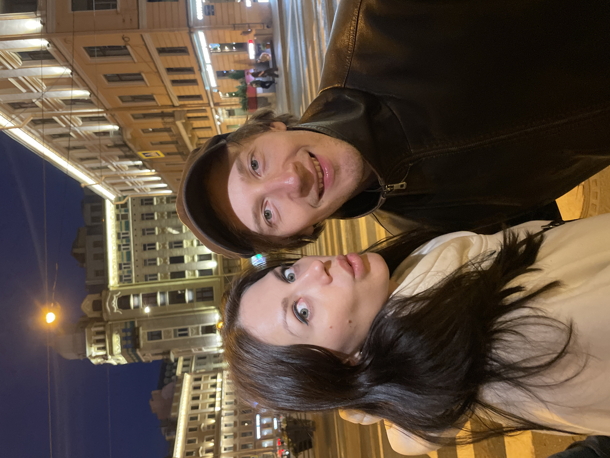 In this medium shot photograph, a man and a woman are positioned in the foreground, posing for a selfie at night on a crosswalk. The man, on the right, wears a brown baseball cap and a dark leather jacket. He has light blue eyes and a wide smile, showing his teeth. The woman, on the left, has long dark hair and a light-colored scarf and coat. She is looking at the camera with wide eyes and her lips pursed.
Behind them, the cityscape of Saint Petersburg, Russia, is illuminated by streetlights and lights from buildings. The architecture suggests a European city with ornate facades and multiple stories. The dark sky indicates it is nighttime. The crosswalk markings are visible on the street, indicating a busy urban intersection. The presence of streetlights and building lights creates a warm, artificial glow over the scene. The overall impression is of a couple enjoying a night out in a vibrant city.
