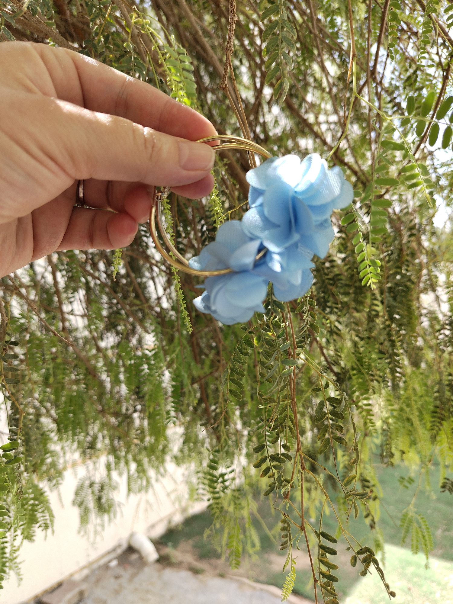 In Al Mas'oudi, United Arab Emirates, a hand holds a pair of gold hoop earrings adorned with light blue fabric flowers. The background features foliage of a tree with small, fern-like leaves. The scene appears to be outdoors, with diffused sunlight and a partial view of a building or structure in the lower portion of the image.