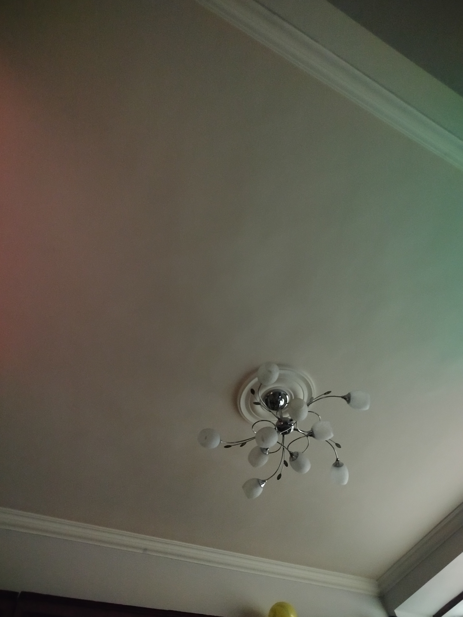 The image shows a ceiling with a chandelier in a room, potentially located in Yerevan, Armenia. The ceiling is white with decorative molding around the perimeter. The chandelier is silver-colored with multiple frosted glass shades, arranged in a semi-random, branching pattern. Below the ceiling, there is a partial view of a wall or other structure, including a glimpse of a yellow object, potentially a balloon. The lighting in the room appears uneven, with a gradient effect across the ceiling.