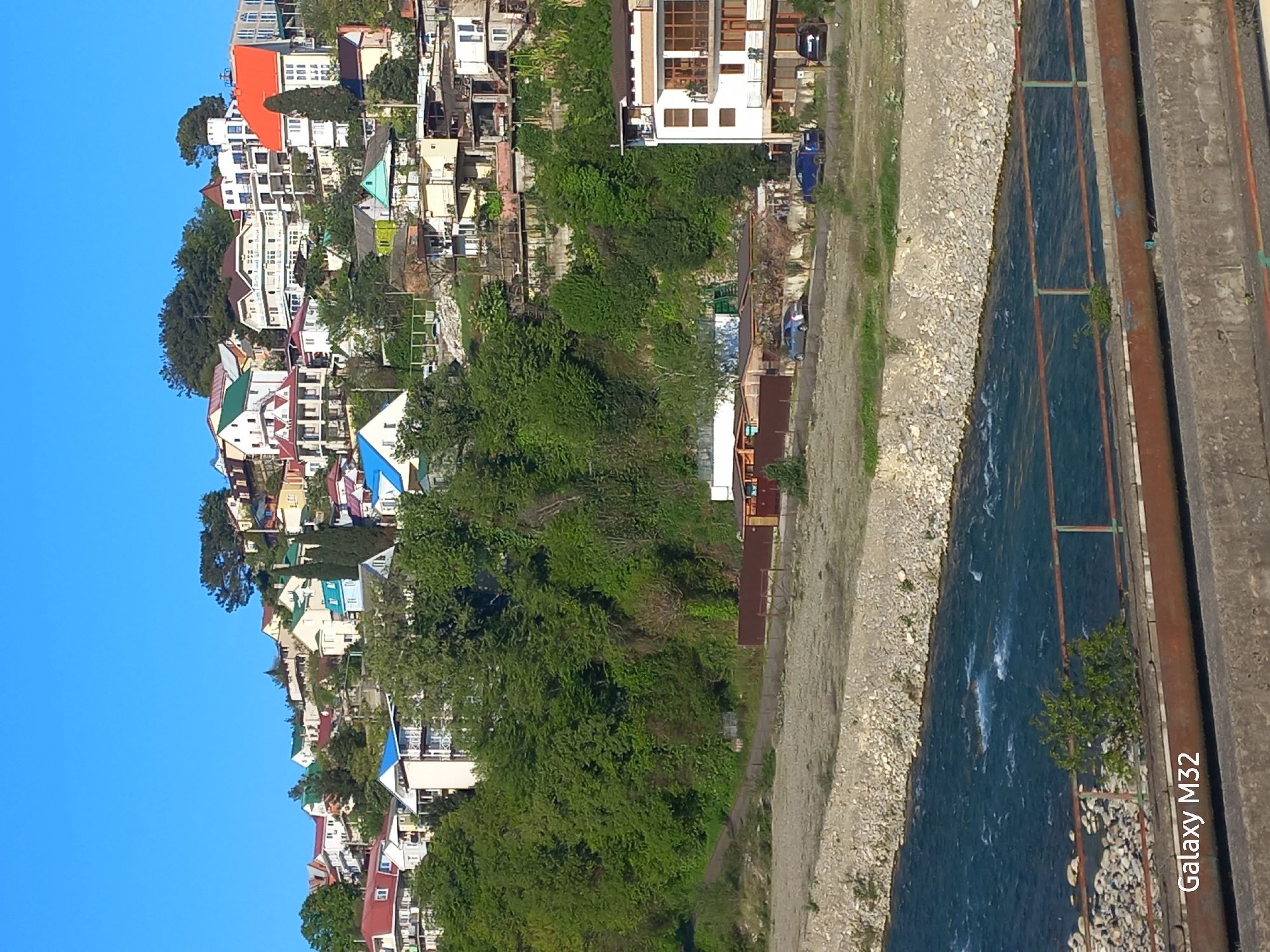The image shows a view in Sochi, Russia, depicting a settlement built on a hillside. A river or canal runs alongside the base of the settlement. The buildings are varied in color and style, featuring roofs in red, blue, green, and other hues. Dense green vegetation covers the hillside and borders the riverbank. On the right side of the frame, there's infrastructure, possibly a bridge or reinforcement structure along the water. The sky is clear and blue.