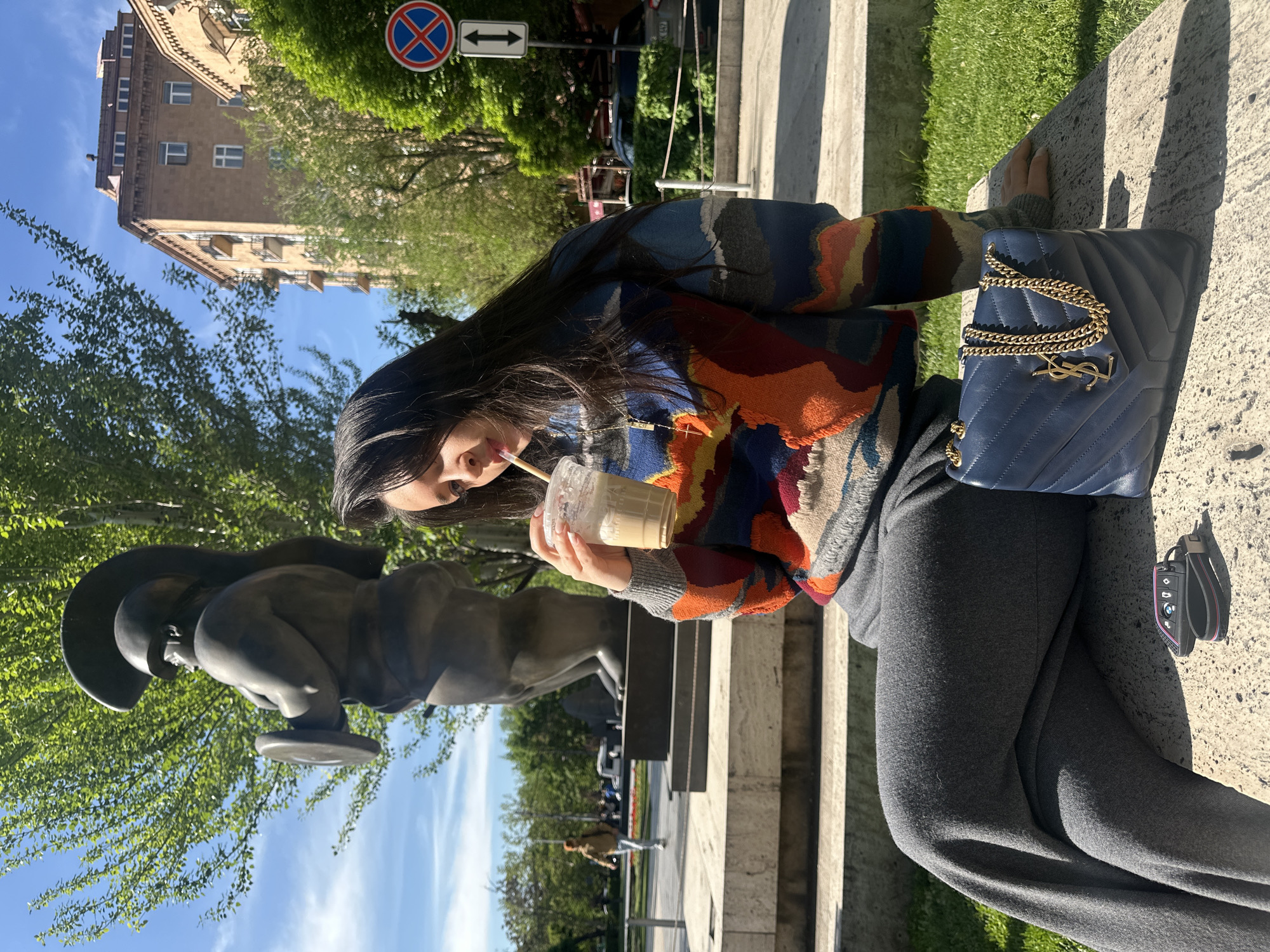 A woman is sitting in Yerevan, Armenia, drinking a beverage through a straw. She is seated on a concrete structure next to a bronze sculpture. A navy blue purse with a gold chain and a gold logo is placed next to her on the concrete. She is wearing a colorful sweater and grey pants. A building, trees, and a street sign are visible in the background.