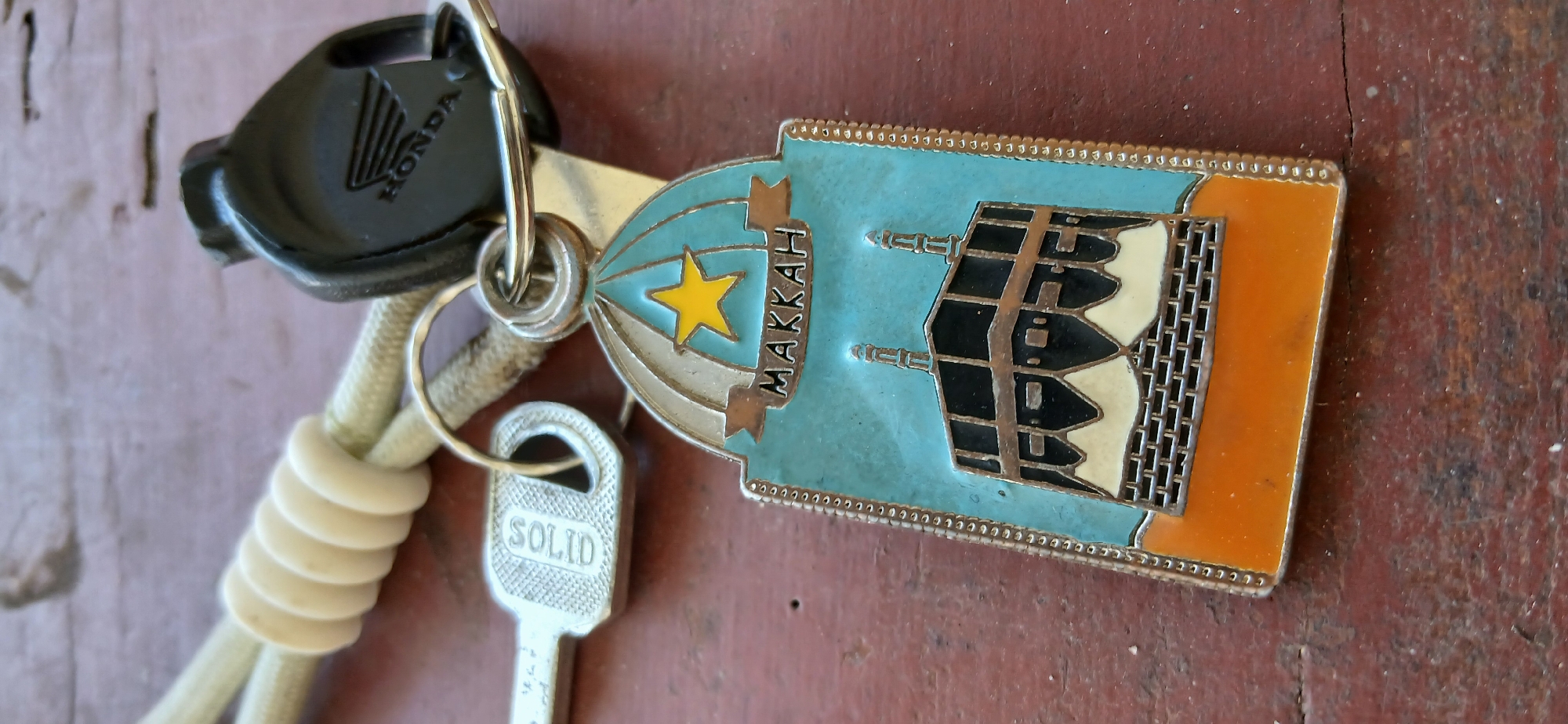 The image shows a close-up of a keychain with a Makkah-themed charm. The charm is a stylized representation of the Kaaba in Mecca, with two minarets and a yellow star above the word "MAKKAH". The charm is attached to a keyring with a Honda motorcycle key. A silver-colored house key with the word "SOLID" is also visible. The items are resting on a wooden surface with a reddish-brown hue and visible grain. The background is blurred, suggesting a shallow depth of field. There are no people or animals in the image. The overall scene appears to be a still life composition.