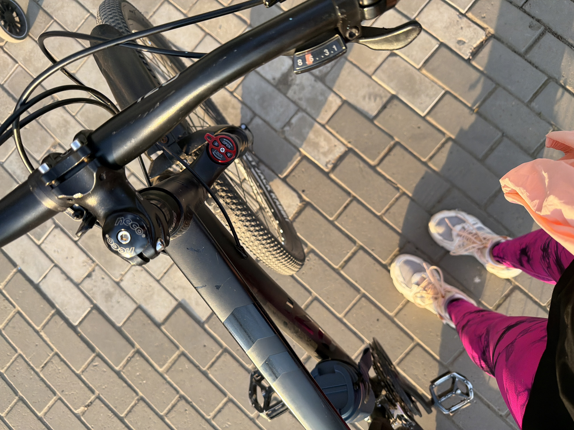 The image shows a bicycle standing on a paved surface, likely a sidewalk or pedestrian area. The bike has black handlebars, a suspension fork with a red lock/open dial, and shifters showing gears. A person wearing pink leggings, white sneakers, and a peach-colored top is standing next to the bike. The background consists of paving stones arranged in a herringbone pattern. Based on the information in the prompt, the location is likely in Chaikovsky, Russia, but this cannot be confirmed from the image alone.