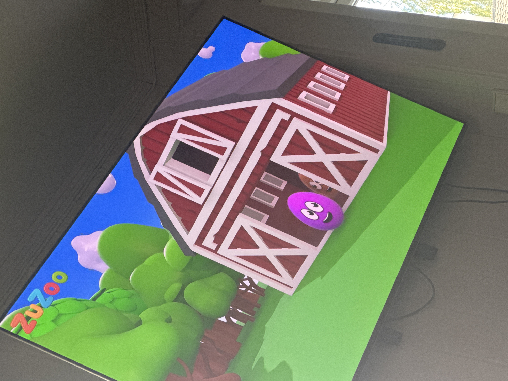 A television screen displays a cartoon scene with a red barn and green rolling hills under a blue sky. A purple, oval-shaped character with a smiling face is in the foreground. The logo "ZuZoo" is visible in the top left corner of the screen. The television is mounted on a wall in what appears to be a living room, with cables visible below the screen. The image is slightly tilted. The television is located in Moelv, Norway, in a residential area.