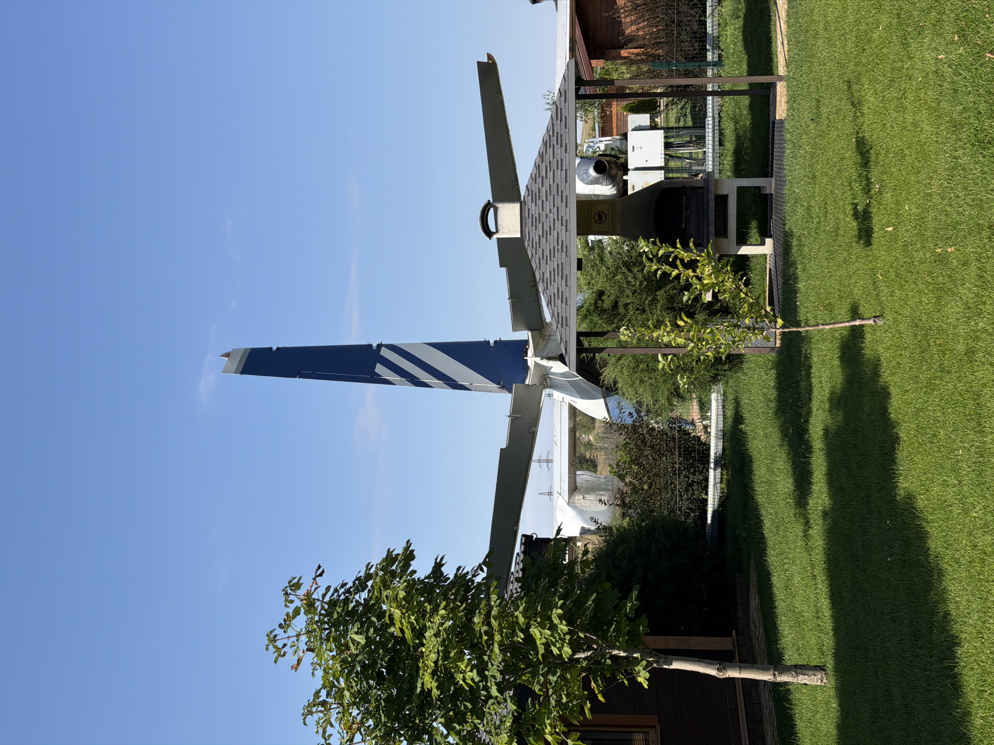 The image captures an outdoor scene featuring the tail section of an airplane. The plane's tail is painted blue with white stripes. In the foreground, there is a manicured lawn and a small tree on the left. To the right of the tree, a barbecue grill is situated under a gazebo with a gray shingled roof. Behind the grill, parts of an airplane are visible, including a wing and an engine. Two small wooden buildings, possibly sheds or guest houses, are present on the left and right sides of the frame. The background consists of trees and a clear blue sky. The location is identified as being in the city of Kamyshin, Russia.