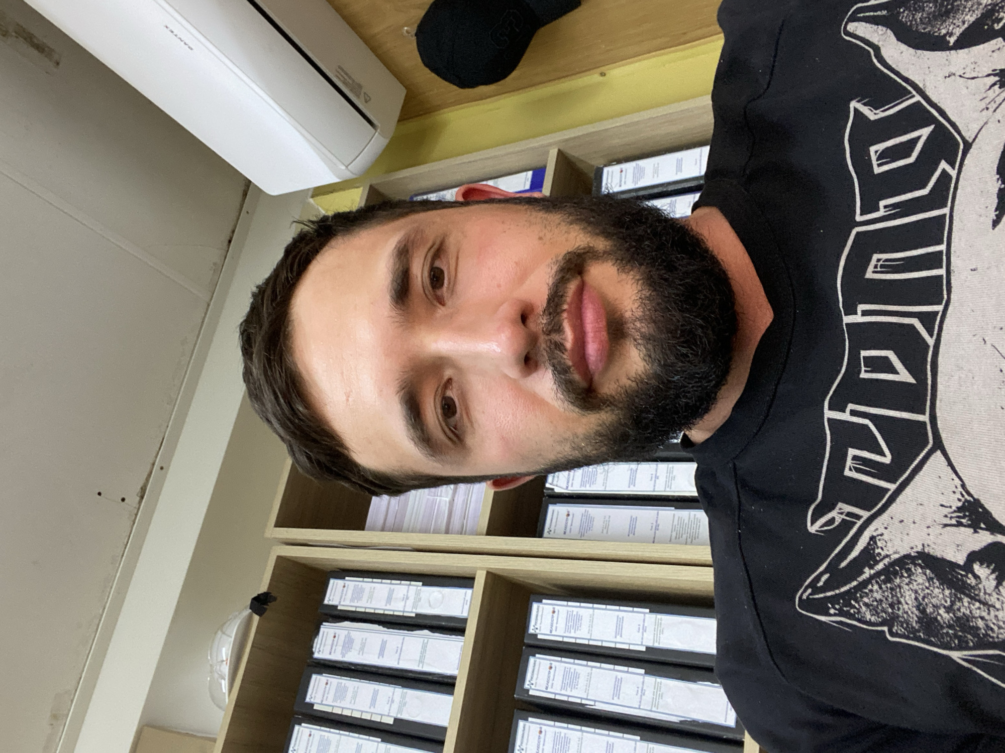 The image is a close-up selfie taken by a man with dark hair and a beard. He is wearing a black t-shirt with a graphic print. He is indoors, with shelves filled with binders behind him. An air conditioning unit is visible on the wall above the shelves. There is also a black hat hanging on the wall. The setting appears to be an office or storage area. The image does not contain any information indicating the location in the city of Luzhitsy, Russia.