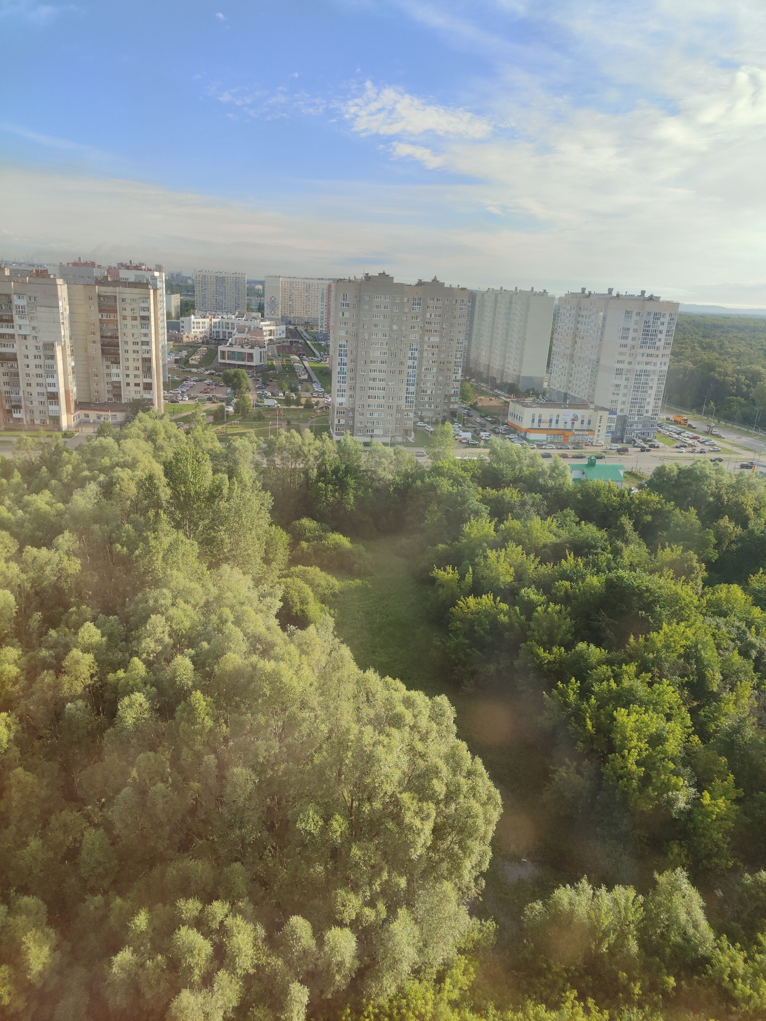 This is a high-angle, vertical photograph of an urban landscape bordering a natural green space. In the foreground and mid-ground, a dense canopy of deciduous trees, predominantly in various shades of green, fills the frame, with some trees appearing taller and fuller than others. The foliage suggests a warm season, likely summer.
Beyond the trees, a city skyline emerges, characterized by numerous apartment buildings. These structures are modern, tall, and appear to be made of concrete and glass, with many visible windows and balconies. Some of the buildings are lighter in color, while others have a grayish hue. In between the residential towers, smaller, more contemporary buildings, possibly commercial or civic structures, are visible.
A road runs alongside the cityscape and the treeline, with cars parked along its edges and moving traffic on the road itself. There is a hint of a traffic signal or street light. A small green-roofed building is also visible near the road. The overall scene suggests a planned community where urban development has integrated with or encroached upon a natural area.
The sky above is a mix of blue and white, with scattered clouds. The lighting suggests it is daytime, possibly late afternoon or early morning, given the soft quality of the light and the shadows. The atmosphere appears clear. There is no discernible text in the image. The scene evokes a sense of everyday urban life juxtaposed with accessible nature.