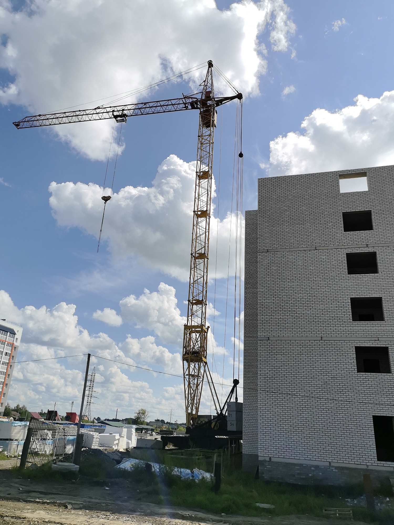 The image shows a construction site in Novoaltaysk, Russia. A tall yellow tower crane is visible, with its boom extending horizontally to the left. To the right of the crane, a partially constructed building made of white bricks is shown. The building has several open window frames on its upper floors. In the foreground, there is a pile of construction materials, including stacks of white insulation panels. The sky is blue with scattered white clouds. A power line tower is visible in the background, along with some residential buildings and trees. The ground is uneven and appears to be dirt and gravel. There are no people visible in the image.