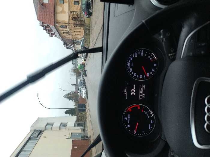 The scene is viewed from the driver's seat of an Audi vehicle, identifiable by the steering wheel logo and dashboard design. The vehicle's dashboard indicates a speed of 33 km/h, with the analog speedometer needle pointing to 30 km/h, and an ambient temperature of +4.5°C. The tachometer shows the engine at idle RPM, approximately 700-800. Windshield wipers are visible in an elevated position, suggesting active use due to rain or mist.
The vehicle is traveling on a wet asphalt road in Saarlouis, Germany, specifically on Holtzendorffer Straße, heading in a southeasterly direction.
On the left side of the road, a large, multi-story, cream-colored commercial or industrial building with a flat roof, multiple windows, and a large ground-level roll-up door is visible. A wooden fence or hoarding separates the building from the sidewalk, with a bicycle parked against it. A modern street lamp with a curved arm stands further down the street.
On the right side of the road, a row of older, multi-story buildings with distinct red-tiled gabled roofs and dormer windows are present. The building closest to the foreground on this side features a light-colored, somewhat worn or patched facade. Several vehicles are parked along the curb on this side of the street.
Further ahead on the road, multiple vehicles are visible, including a dark sedan directly in front of the camera vehicle, a white van on the left side of the street, and a green truck or bus further down on the right. The sky is overcast, indicating dull, diffuse lighting conditions.