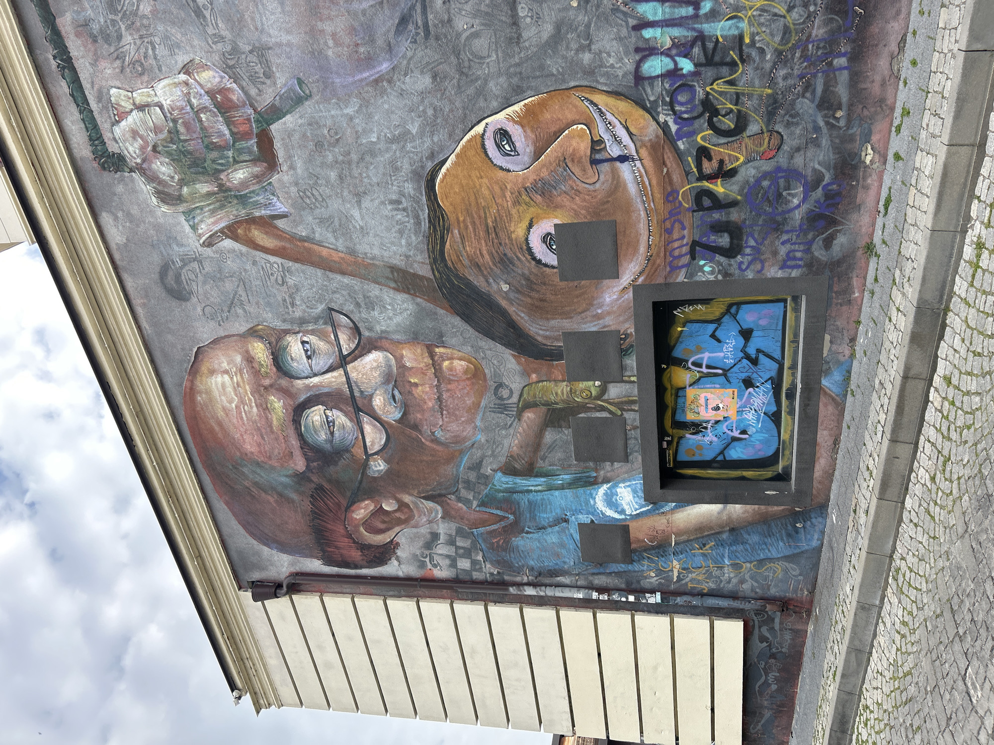 A mural is painted on the side of a building. The mural has two large human faces, one wearing glasses and the other holding a rope or cable in a gloved hand. A framed opening in the wall is filled with graffiti. The building abuts a cobblestone street. The mural is in Plovdiv, Bulgaria.