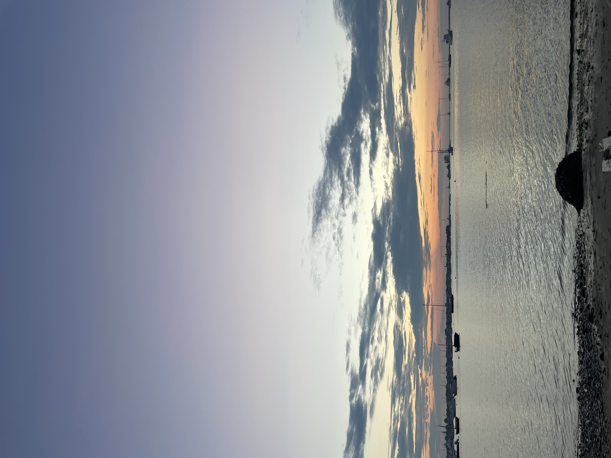 This image depicts a serene harbor scene at dusk in Nantucket, United States of America. The sky transitions from a soft lavender at the top to hues of pale orange and pink near the horizon, where scattered clouds are visible. The water, reflecting the muted colors of the sky, is calm with gentle ripples. Several sailboats and motorboats are anchored or moored in the harbor, silhouetted against the twilight sky. In the foreground, a pebble-strewn shoreline leads to the water's edge, with a dark, mossy object resting on the sand. The overall atmosphere is tranquil, capturing the quiet beauty of a coastal evening.