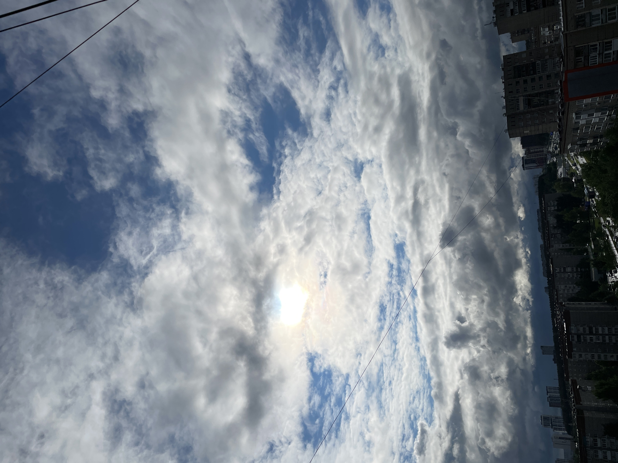 The image depicts a view looking upwards in the city of Izhevsk, Russia. A bright sun is visible through a sky filled with layered, textured clouds, ranging from white to gray. High-rise apartment buildings line the upper right of the frame. Electrical wires extend across the sky. Street trees are visible beside the buildings.