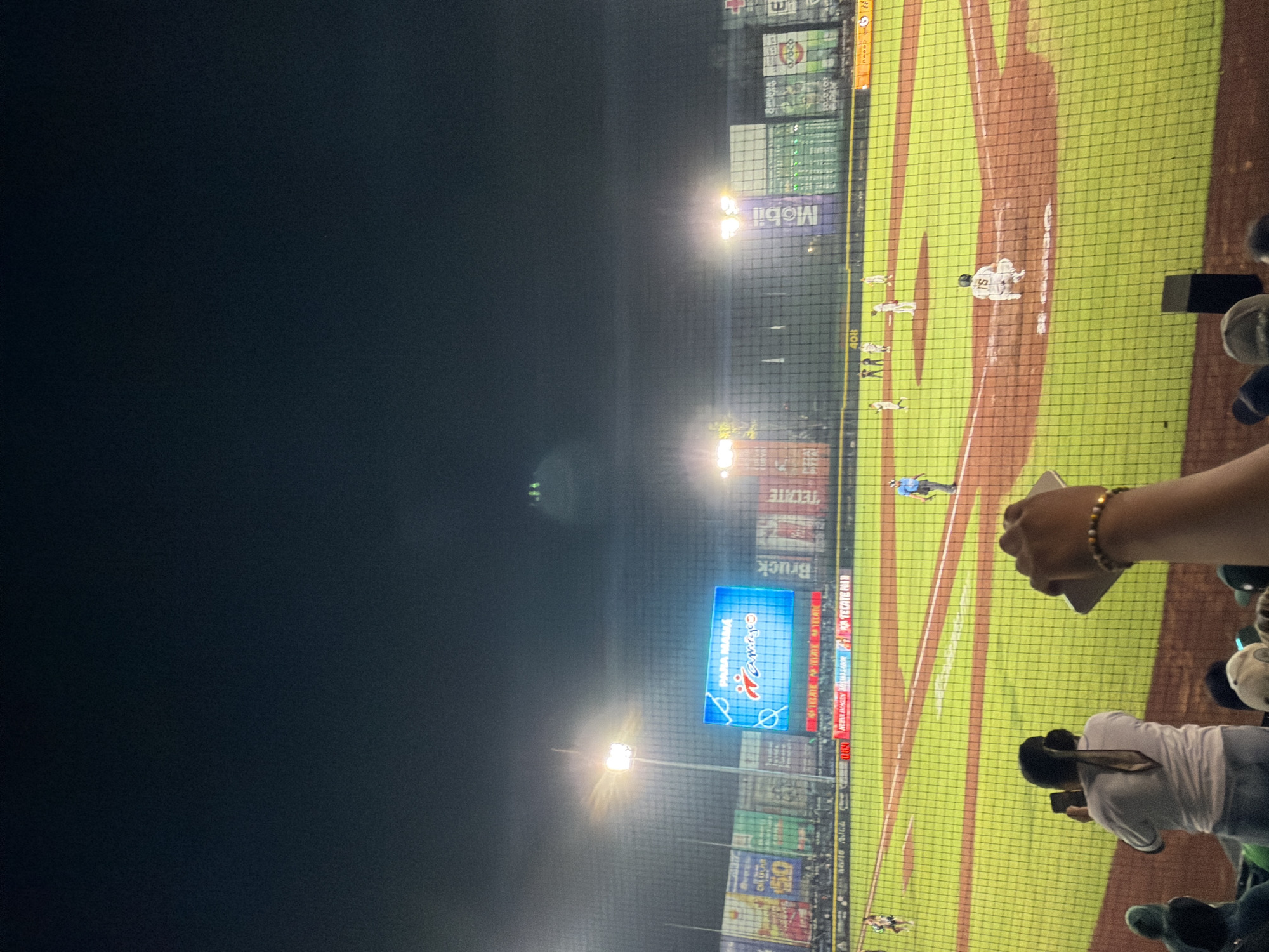 The image depicts a baseball game at night, likely taken from stadium seating behind a protective net. The field is illuminated by stadium lights, and several players are visible on the field wearing white uniforms. A large scoreboard displays advertisements, including one for "Para Llevar Jumex" (which indicates that the game is most likely being played in Mexico since Jumex is a very popular Mexican Juice Brand), as well as other commercial brands. Spectators are present in the foreground, with at least two individuals visible taking photos or videos with their phones. Considering the ads that were visible on the scoreboard and the fact that "Para Llevar Jumex" is visible, there is a high probability that this picture was taken in Puebla, Mexico.