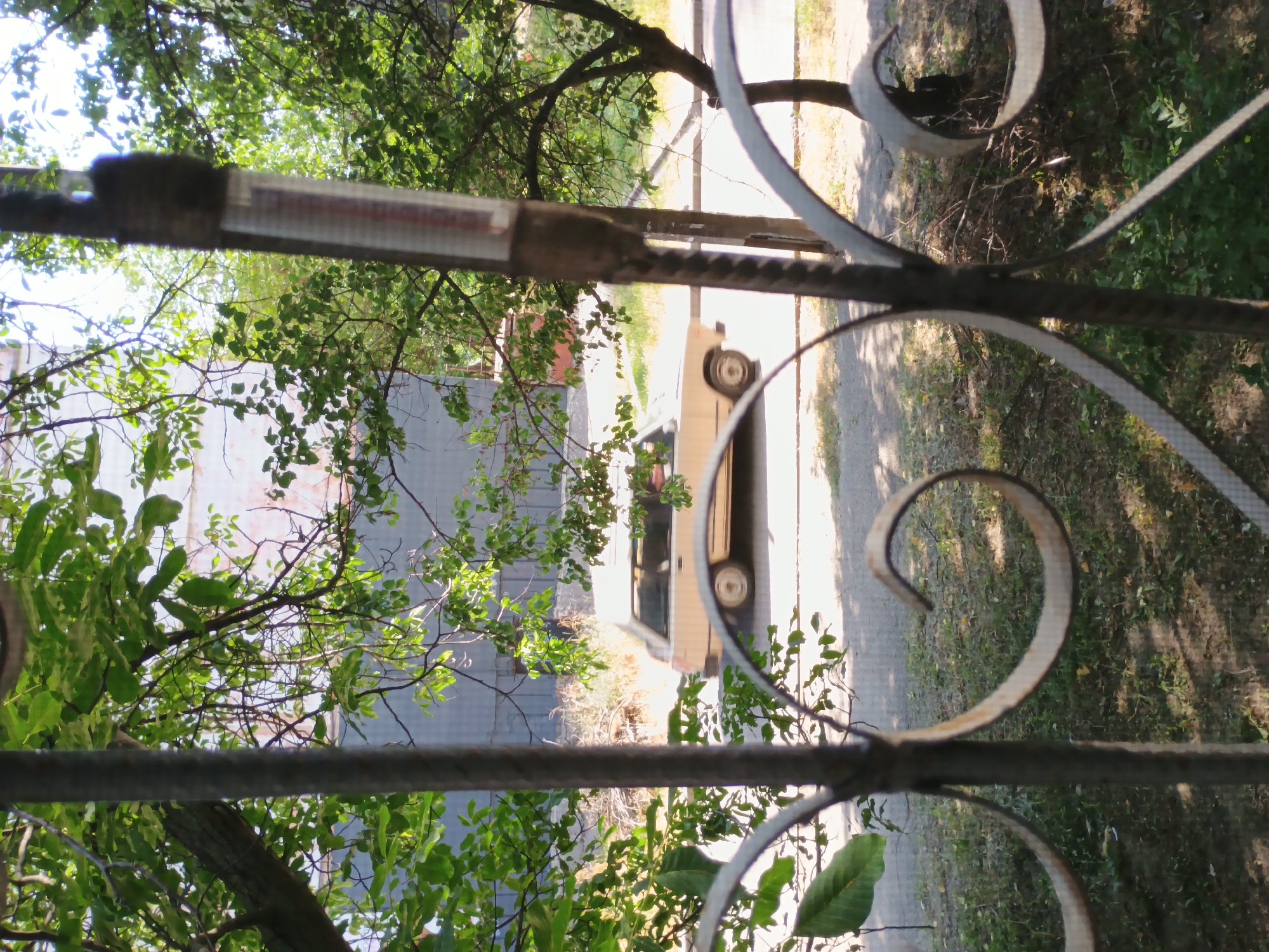 The image shows a view through a decorative metal fence looking out onto a street scene. The fence has a scrolling, ornamental design. Through the fence, a light-colored car is parked on a paved surface, with some greenery and trees in the background. Sunlight filters through the leaves of the trees, creating dappled shadows. The overall impression is of a sunny day. There is some visible text on a piece of tape attached to a horizontal bar of the fence. The image appears to be taken outdoors, likely in a residential or urban area. The location context of Zaporizhzhia, Ukraine, suggests this is a scene from that region. No people are visible in the frame.