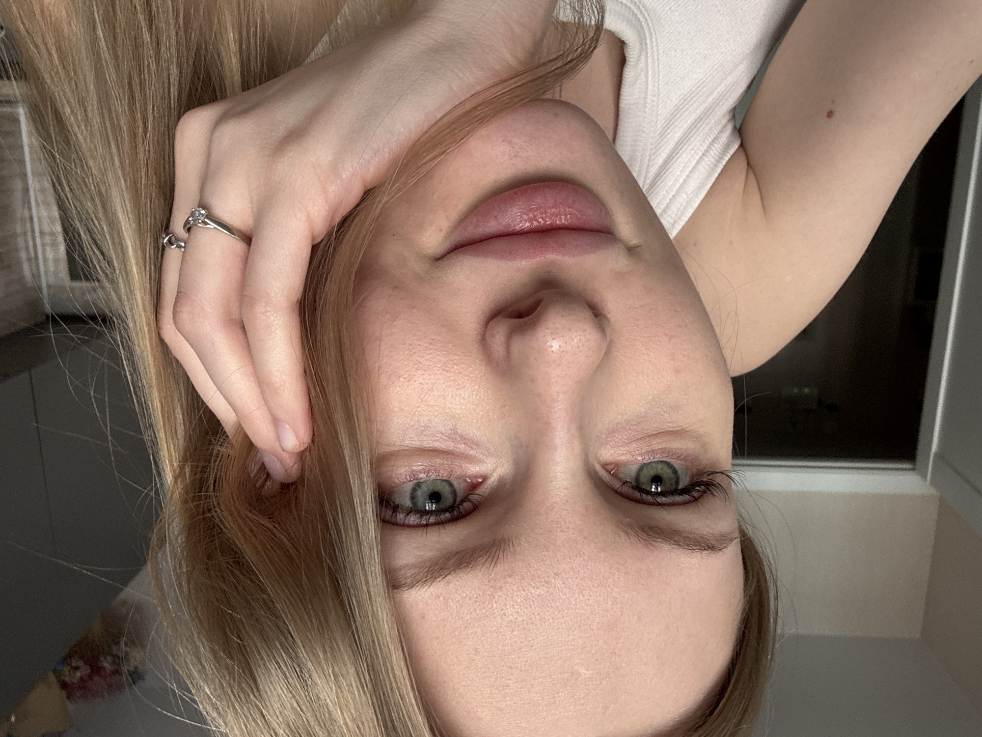 A close-up, inverted portrait features a young woman with long, light blonde hair and greenish-blue eyes. Her face is centered, looking directly at the viewer, with her head appearing upside down due to the image orientation. Her lips are slightly parted, revealing their natural pink hue.
She is wearing a white, ribbed top, visible on her right shoulder and arm. Her left hand is raised, with fingers touching her hair near her forehead, wearing a silver-colored ring with a clear stone on her ring finger. Her fair skin shows a small, reddish-brown mole on her upper right arm.
The background is mostly obscured by her hair, but a dark area with faint, distant lights, possibly from a city at night, is visible through a window on the right side of the frame, suggesting an evening or nighttime setting. No visible text is present. The scene is an indoor setting, likely in Perm, Russia, as per the provided context.