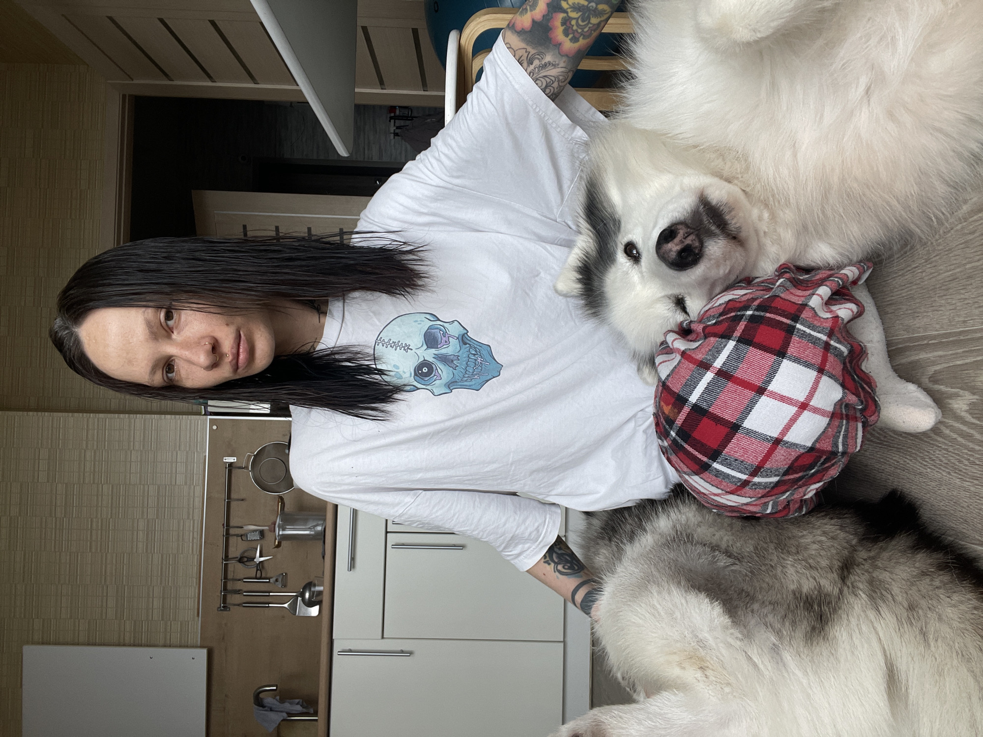 A medium shot shows a woman with wet, dark, straight hair standing in a kitchen. She looks directly at the camera with a neutral expression. She wears a white T-shirt with a light blue skull design. Both of her arms are covered in tattoos. Lying on top of her is a white and grey Siberian Husky dog. The dog is on its back with its head nestled against a red and white plaid ball-shaped object. The background shows a kitchen with beige textured wallpaper. Kitchen utensils are hanging from a metal rack on the wall. White kitchen cabinets with silver handles are visible. Given the lack of specific landmarks or identifiable urban features in the photograph, determining the precise location within the city of Izhevsk, Russia is not possible.