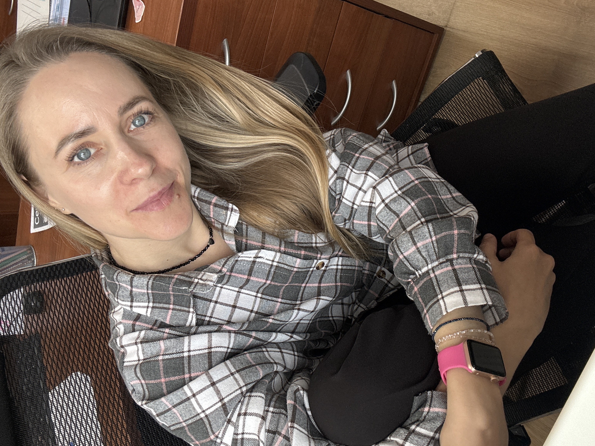A woman with blonde hair and blue eyes sits in a black mesh office chair, wearing a gray, white, and pink plaid shirt, black leggings, and a pink smartwatch. She is indoors, next to a dark brown wooden desk or cabinet. The location is potentially Chaikovsky, Russia.