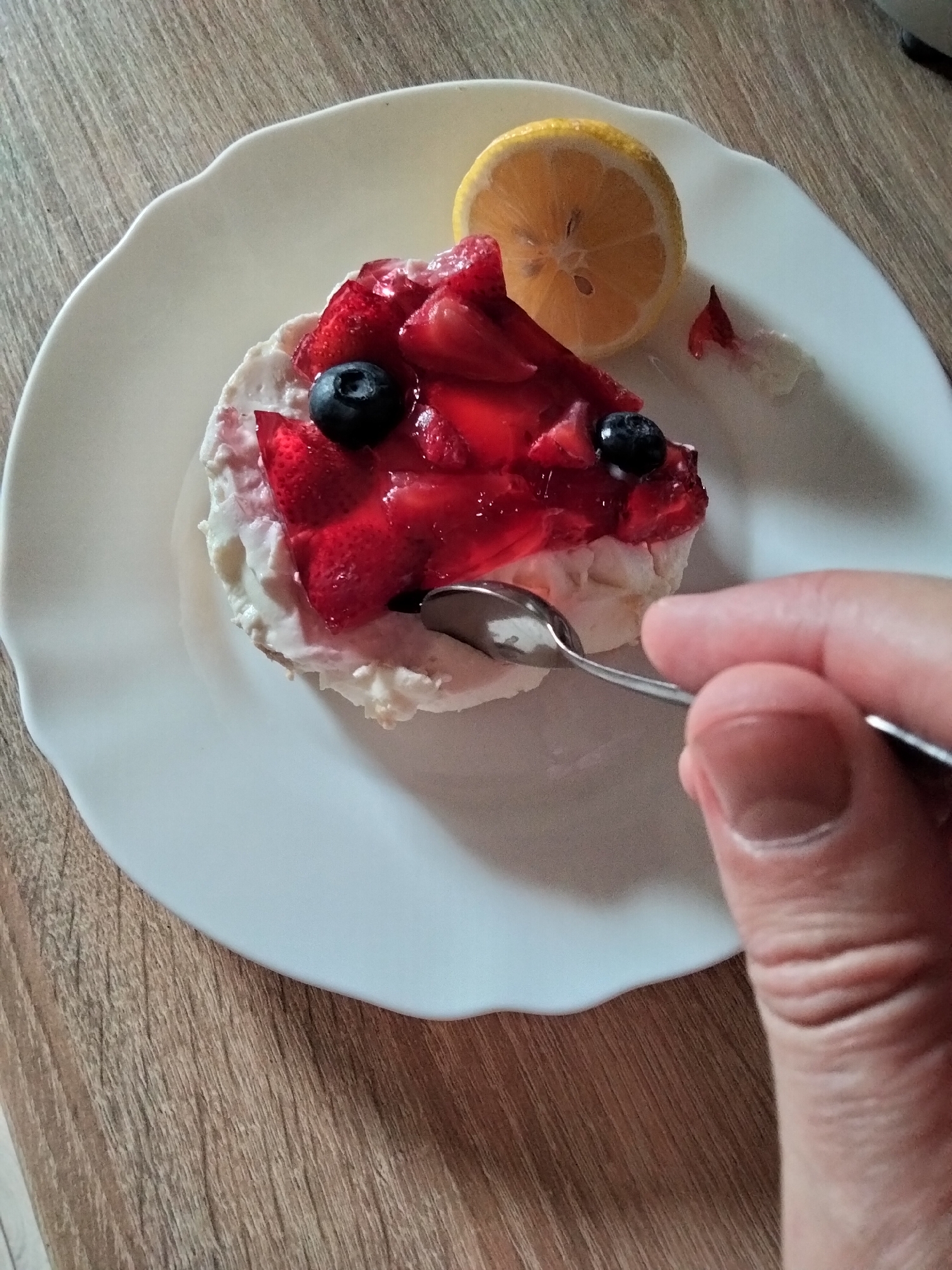 A hand is using a spoon to eat a dessert on a white plate. The dessert is topped with strawberries, blueberries, and jelly. A slice of lemon is on the plate next to the dessert. The plate is on a wooden table. The scene appears to be indoors. There is no information available regarding the location in Kyiv, Ukraine.