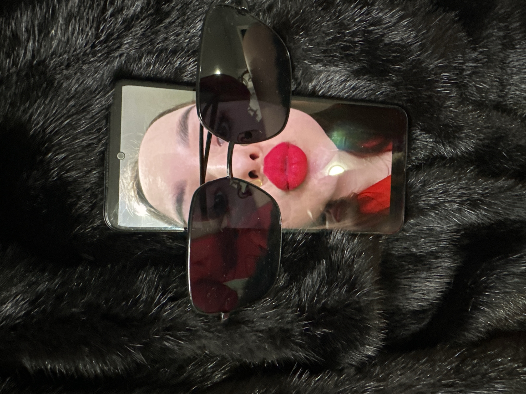 The image shows a pair of sunglasses placed on top of a smartphone. The smartphone displays an image of a woman's face with a prominent red element covering her nose. The background is a dark, textured surface resembling fur. The location is Chaikovsky, Russia.