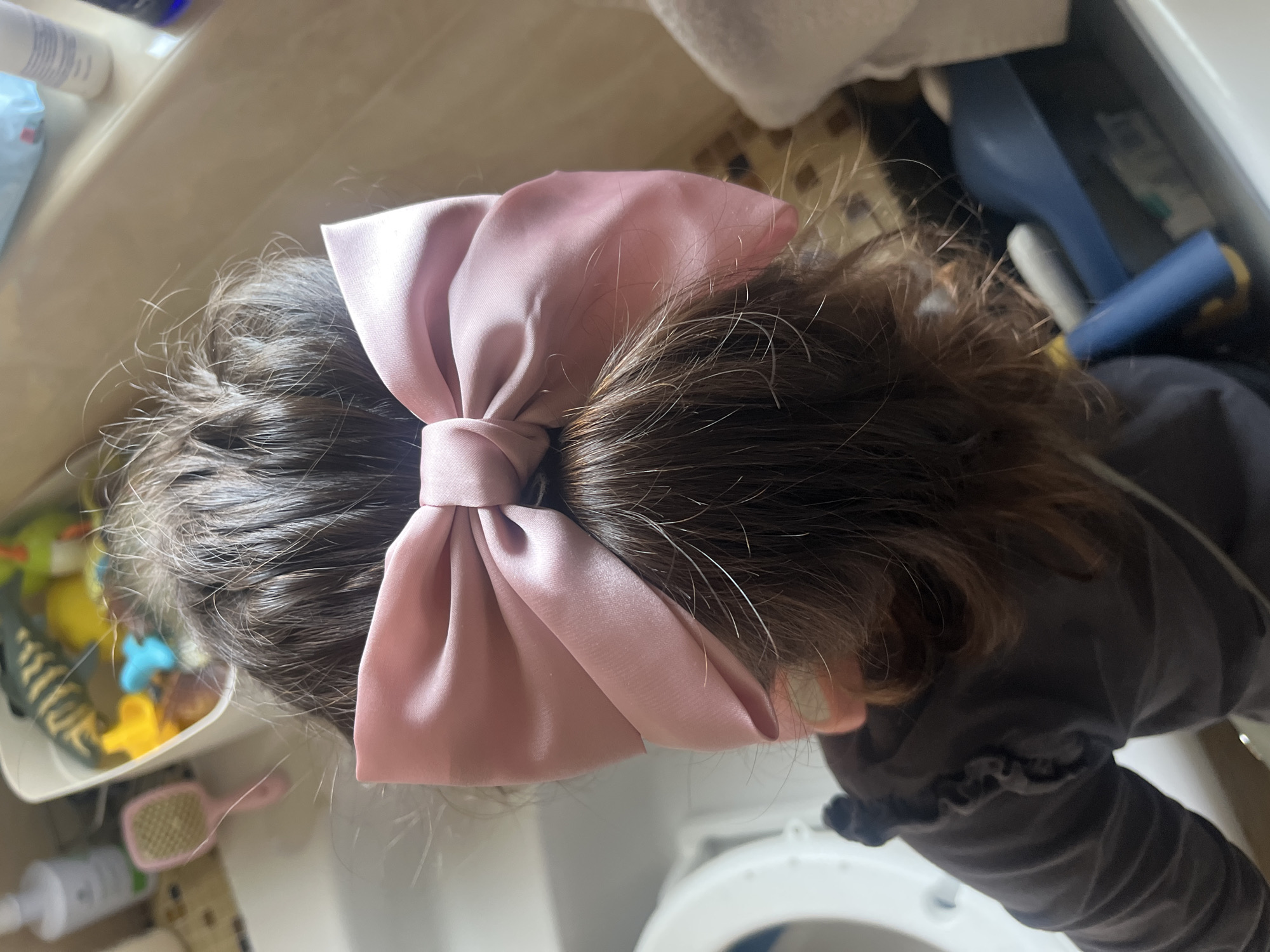 A top-down view reveals a child's head with dark hair, adorned with a large pink satin bow. The child is wearing a dark grey, long-sleeved garment. The background includes a white surface that appears to be part of a bathroom fixture and a white drawer with various items inside, including toiletries, toys, and a pink hairbrush. The location is speculated to be Onokivtsi, Ukraine. There is no visible evidence to confirm this.
