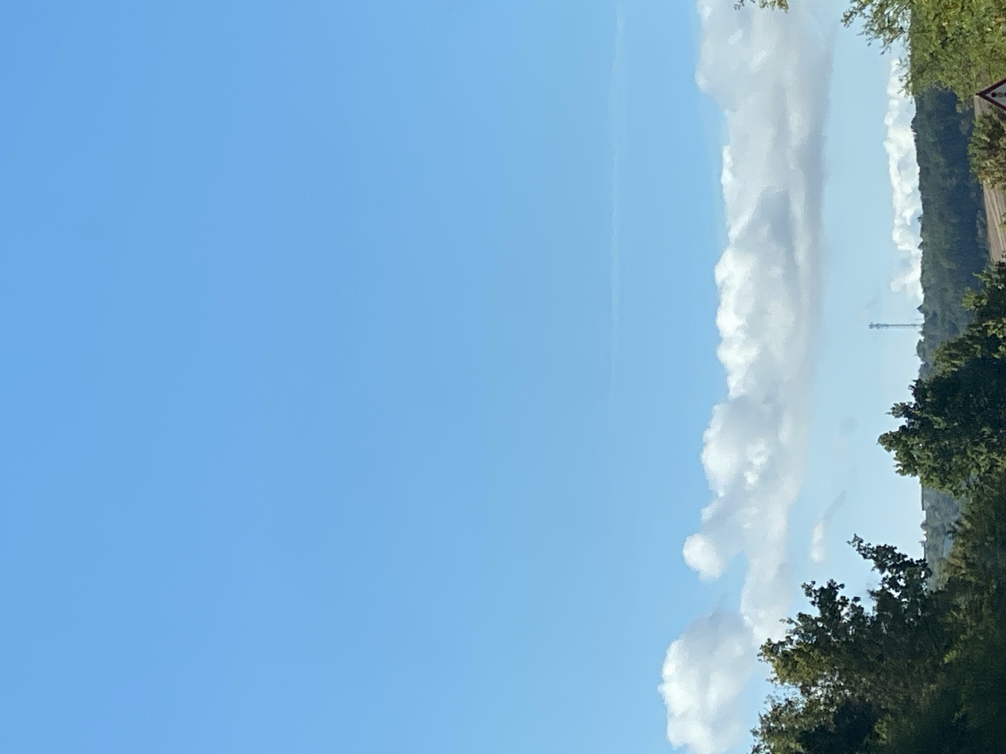 An outdoor, vertical, medium shot captures a clear, bright blue sky dominating the upper portion of the frame. A line of fluffy white clouds stretches horizontally across the middle of the image. Below the clouds, a dense line of dark green trees forms a verdant canopy, with the tops of some taller trees reaching towards the clouds. In the distance, a subtle hill or ridge is visible, dotted with more trees. A slender, tall cell tower stands prominently against the sky above the distant treeline, located on the left side of the frame relative to the distant landscape. A small, triangular warning sign with a red border is partially visible in the lower right corner. The overall impression is of a serene, sunlit day in a rural or semi-rural setting, possibly on the outskirts of Nordhausen, Germany.