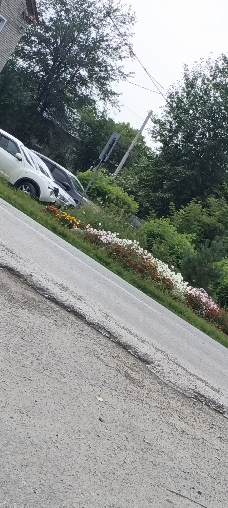 The image depicts an outdoor scene, likely in a suburban or semi-urban setting, with a focus on a roadside area. In the foreground, a cracked asphalt road is visible, angled from the lower left to the upper right of the frame. Beyond the road, a strip of green grass separates it from a flower bed. This flower bed contains a variety of colorful flowers in shades of orange, yellow, white, and pink, arranged in a linear fashion. Behind the flowers, several cars are parked on a grassy incline. The cars include a white SUV and a darker colored vehicle. Further in the background, lush green trees and foliage create a dense natural backdrop. The sky is overcast, suggesting cloudy weather. The overall impression is a roadside scene with a decorative flower border and parked vehicles.