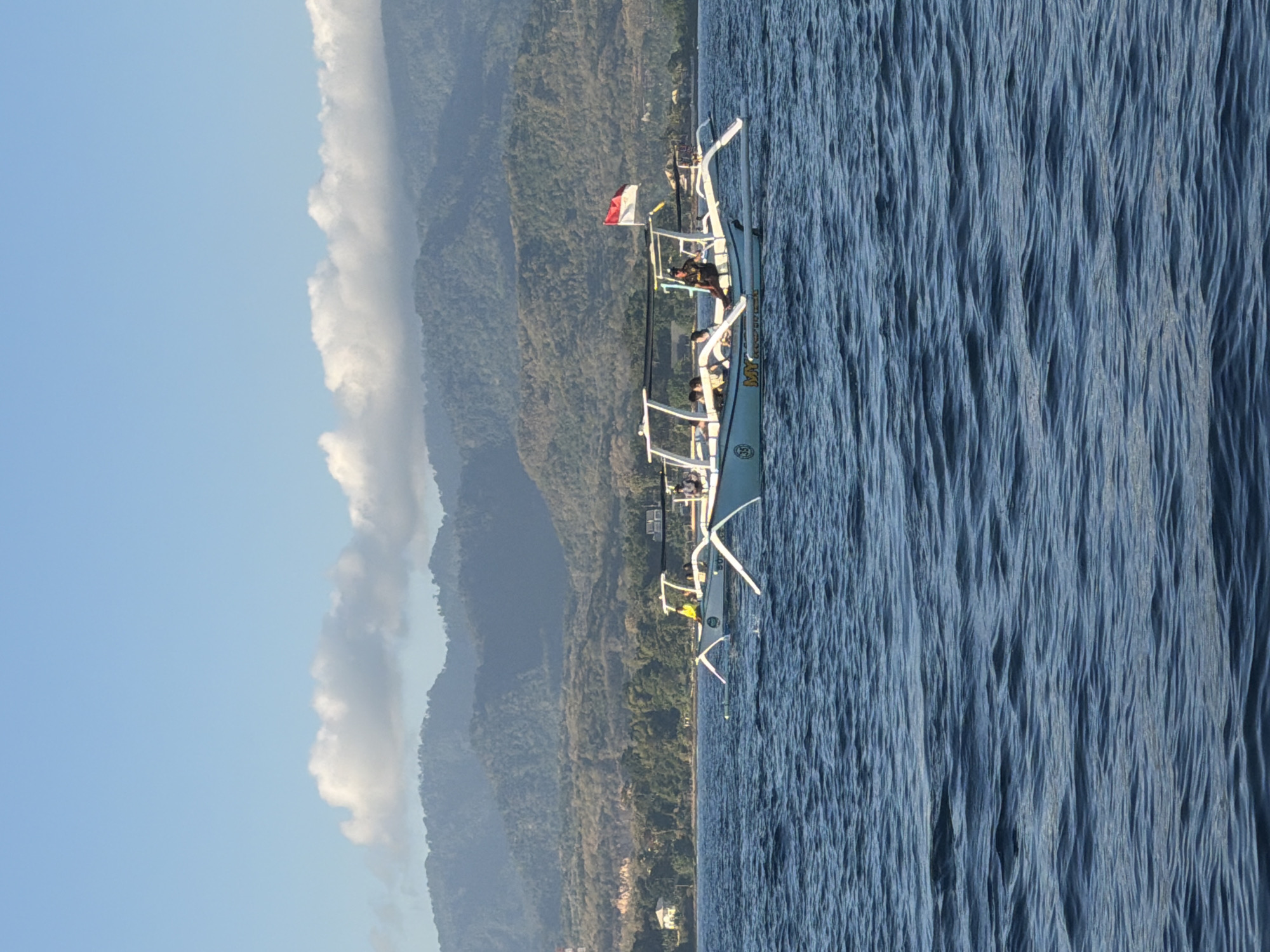The image depicts a traditional Indonesian fishing boat with an outrigger, commonly known as a jukung, sailing on a body of water. The boat is white with blue accents and features a canopy providing shade for its occupants. Several people are visible on the boat, some seated and others standing. A prominent Indonesian flag, red and white, is flying from a pole on the boat.
In the background, a lush, green mountainous landscape stretches across the horizon. The mountains are covered in dense vegetation, and the terrain appears hilly and varied. The sky above is a clear blue, with a few fluffy white clouds scattered across it. The water is a deep blue, with visible ripples and waves suggesting a calm but active sea. The overall scene evokes a sense of a typical day at sea in a tropical coastal region of Indonesia.