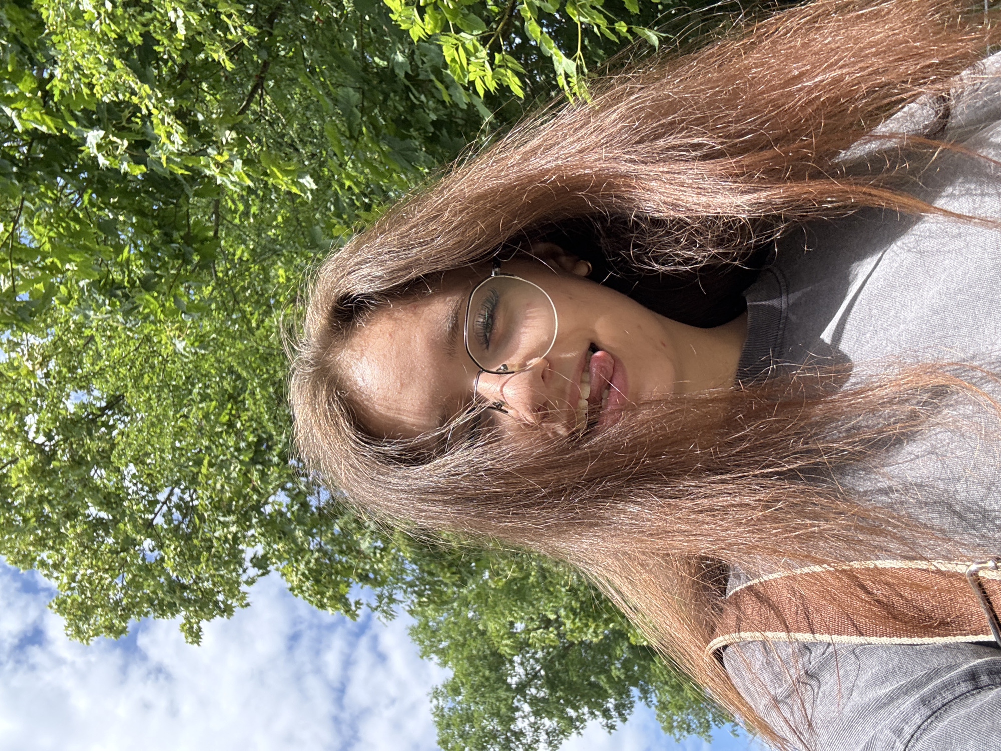 A young person with long, reddish-brown hair, wearing round glasses and a grey t-shirt, is captured in a selfie taken outdoors. They are sticking their tongue out, with their head tilted slightly. Behind them, lush green trees are visible against a bright blue sky with scattered white clouds. The image is framed from the chest up, suggesting a casual, perhaps spontaneous, moment. The natural lighting and the background indicate a sunny day in a park or a similar green space, possibly within the city of Göttingen, Germany, given the context of the prompt. A brown woven strap from a bag is visible on the person's left shoulder.