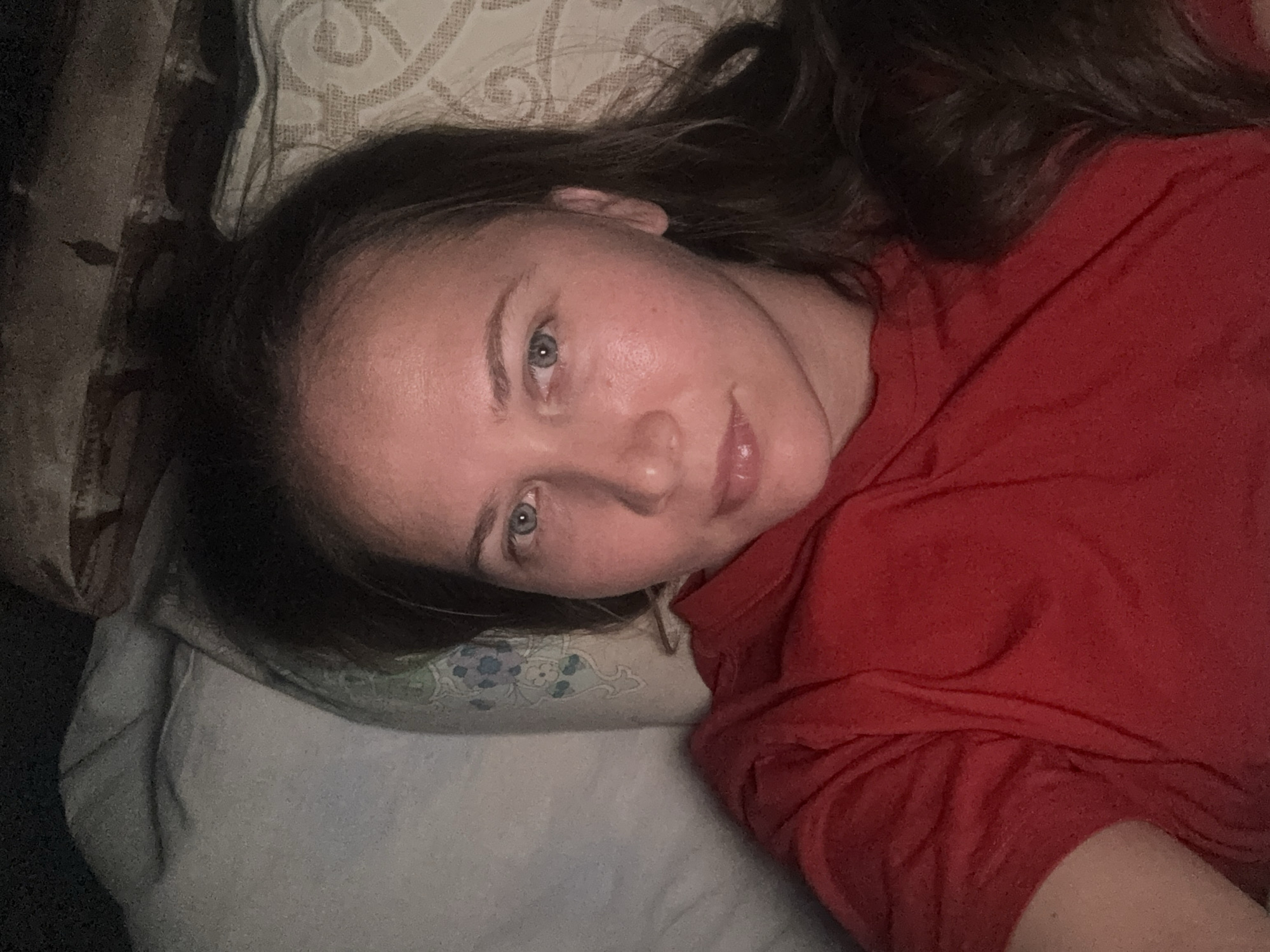 A woman with light skin, blue eyes, and dark brown hair is lying on a pillow. She is wearing a red t-shirt. The pillow has a light-colored patterned case with subtle floral accents. The background is out of focus, but a dark-colored, possibly upholstered surface is visible to the left. The location is not discernible from the image.