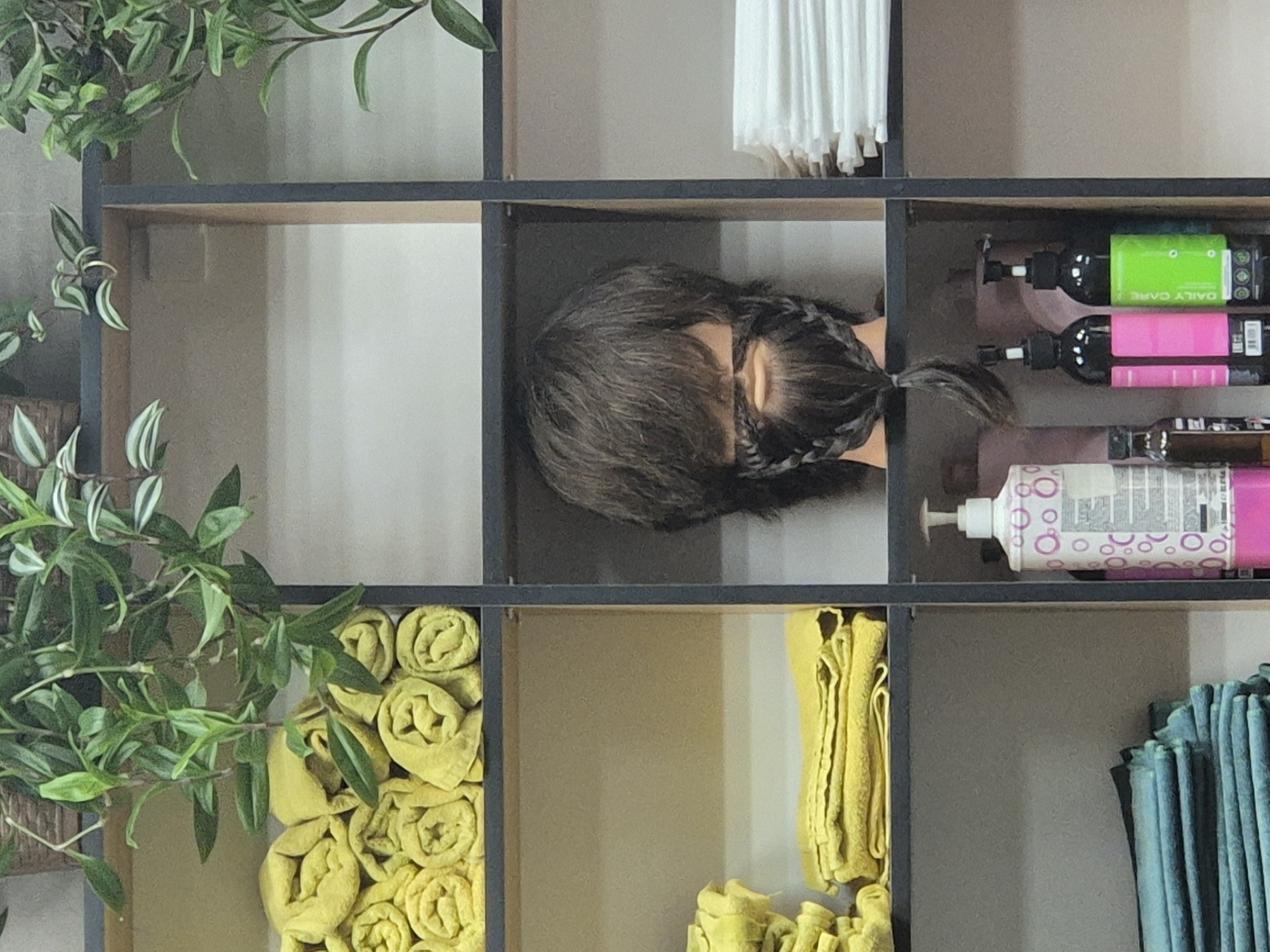 The image displays a set of shelves in what appears to be a salon or barbershop. On the top shelf, several rolled yellow towels are neatly arranged. Below this, on the left side, more yellow towels are folded. The center shelf features a mannequin head with dark, shaggy hair and a braided beard and mustache. To the right of the mannequin, a stack of white folded items, likely towels or linens, is visible. The bottom shelf on the right holds two pump bottles, one with a pink label and another with a green label, and a smaller dark bottle. On the bottom left, a stack of folded teal-colored towels is present. Lush green plants are seen cascading from the top, suggesting a decorative element in the establishment. The overall environment appears clean and organized, typical of a professional grooming service. The location is identified as Kokshetau, Kazakhstan.